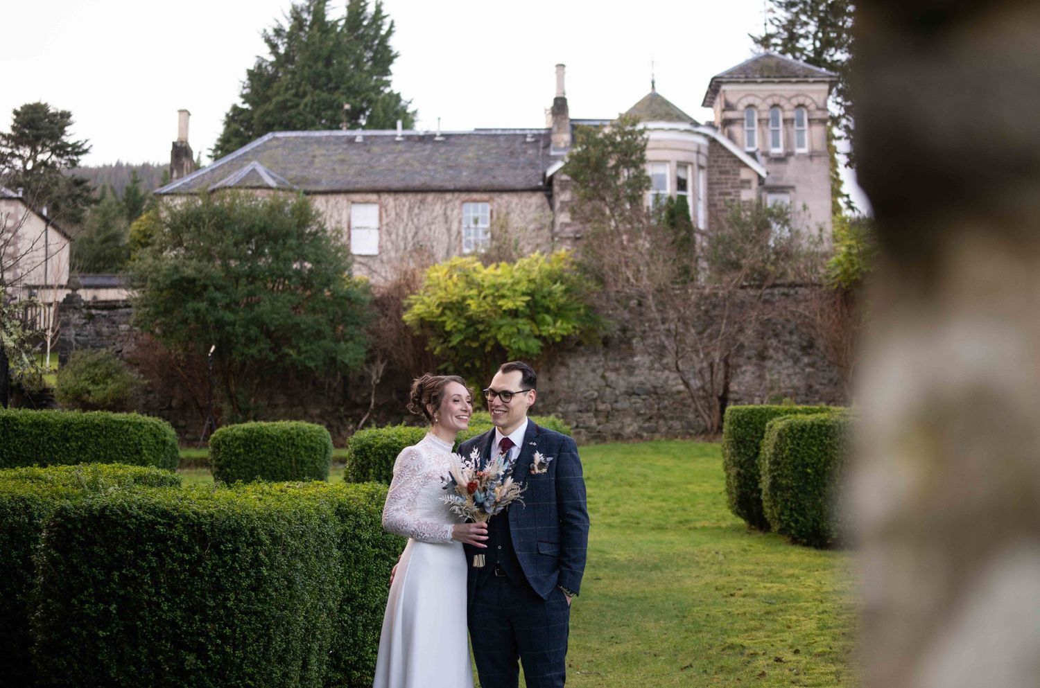 Couple shares romantic moments in a garden setting with stone buildings and hedges in the background.