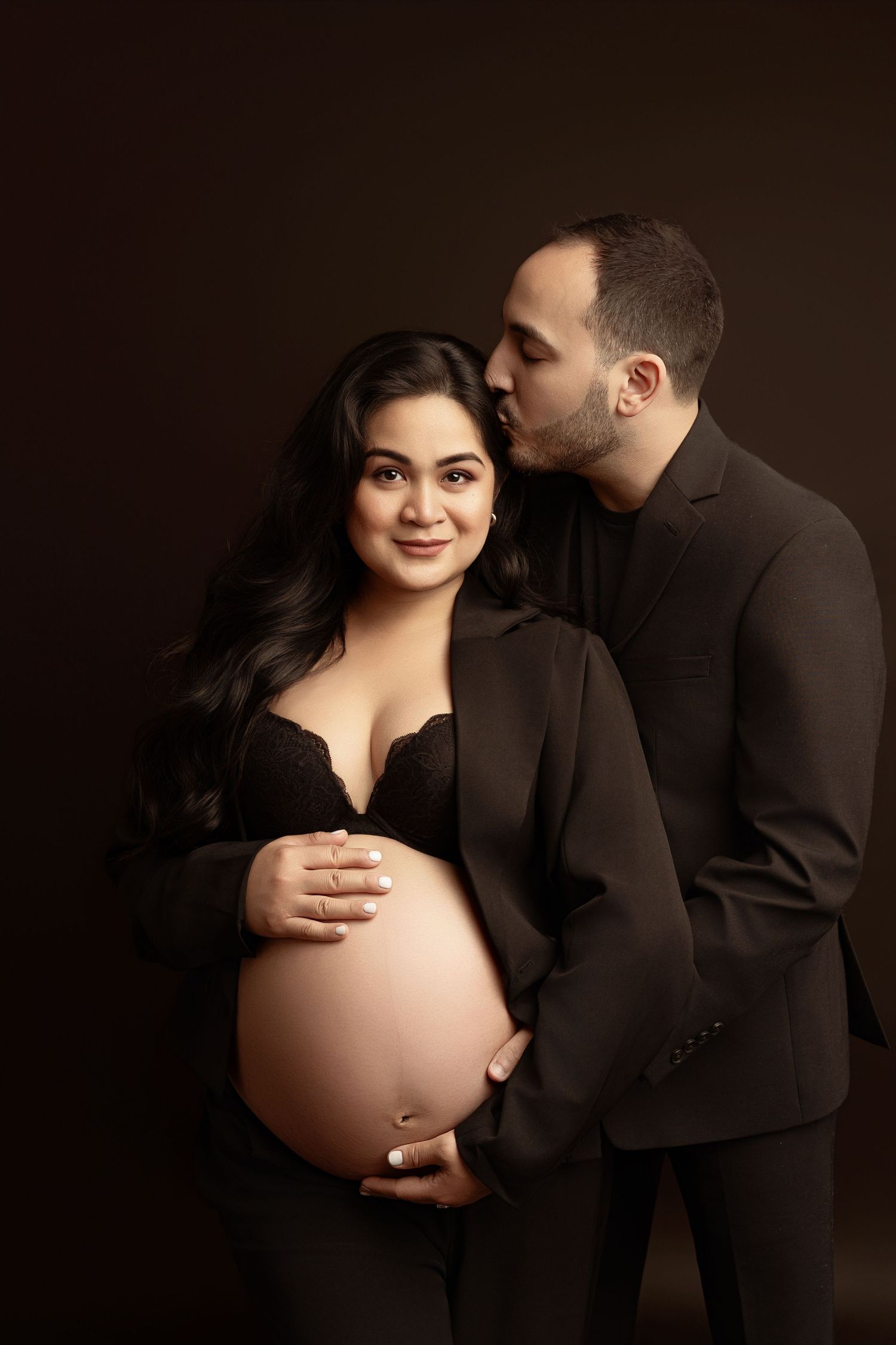 Intimate maternity photo of couple in casual attire embracing against neutral background, showing tattoos and baby bump. Paloma Schell