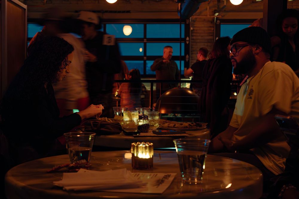 Diners seated at tables in a dimly lit restaurant with large windows at dusk.