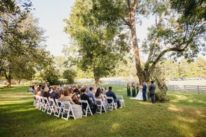 Outdoor wedding ceremony setup with white chairs under large trees.