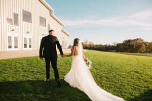 Wedding couple walking across green hillside with white house in background.