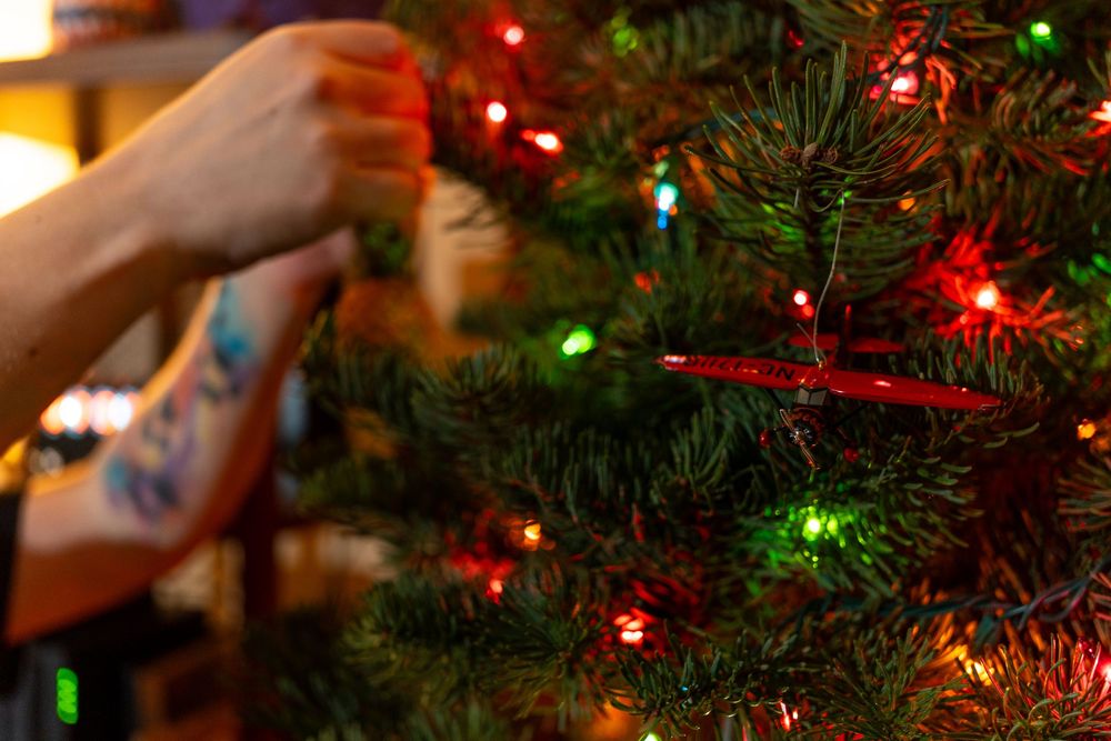 A hand reaches up to hang a shiny red Christmas ornament on an illuminated evergreen tree with colorful lights.