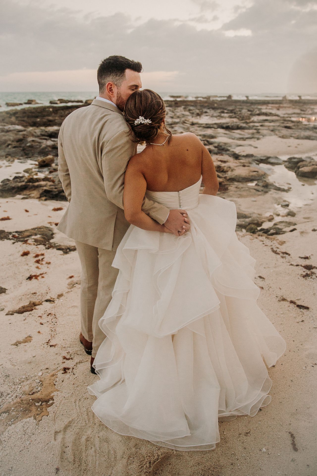 A romantic beach wedding photo shows couple embracing on rocky shoreline with bride in flowing white gown at  El Dorado Royale Wedding Photo