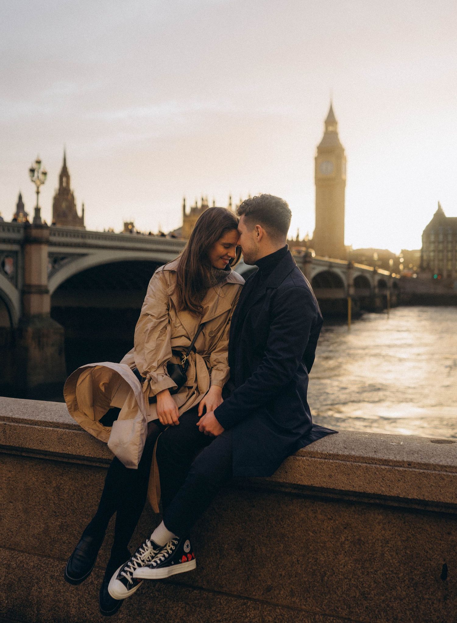 A couple shares romantic moments along a riverside with London landmarks in the background.
