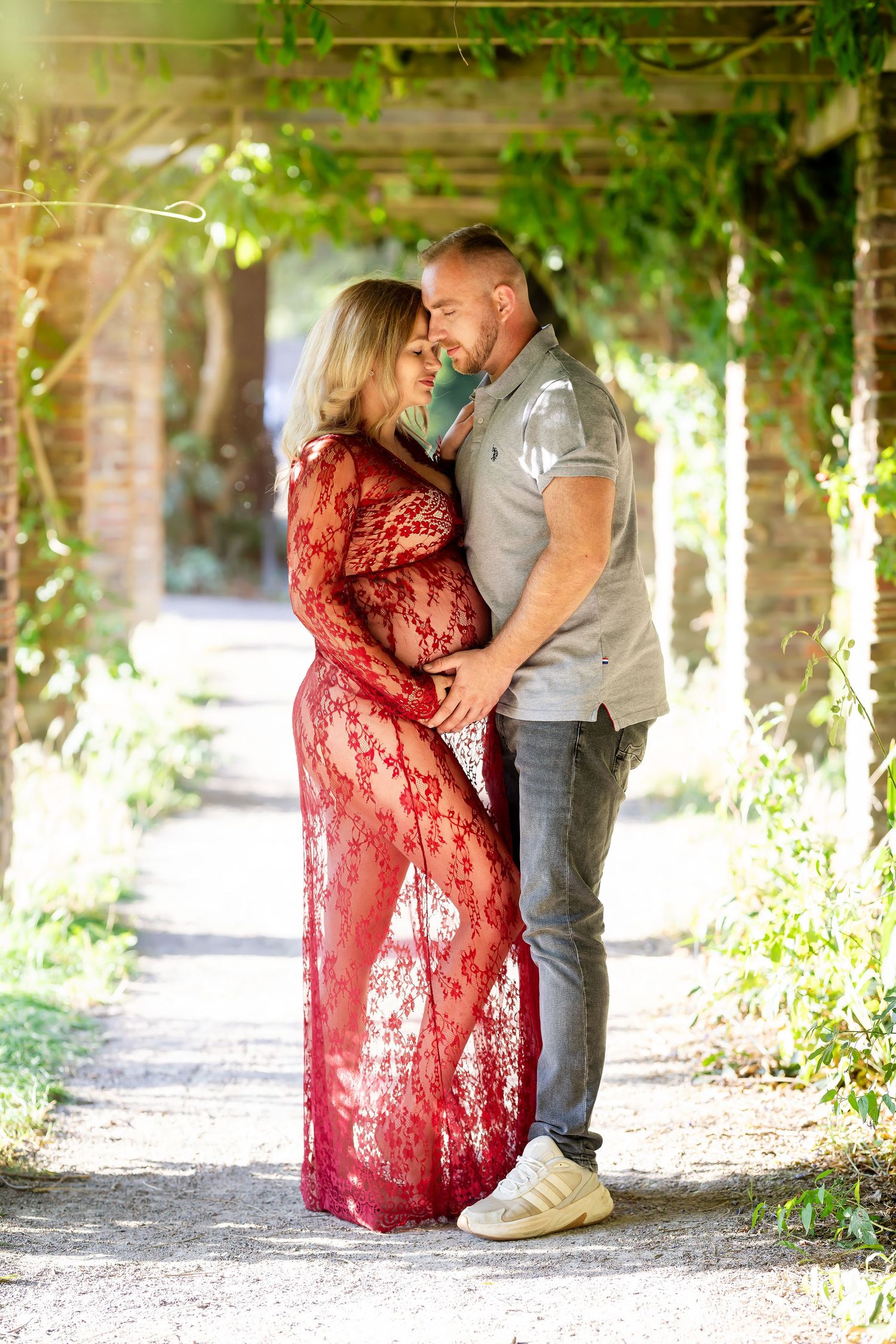 A romantic couple embraces in a garden pathway with green vines overhead and sunlight filtering through the foliage.