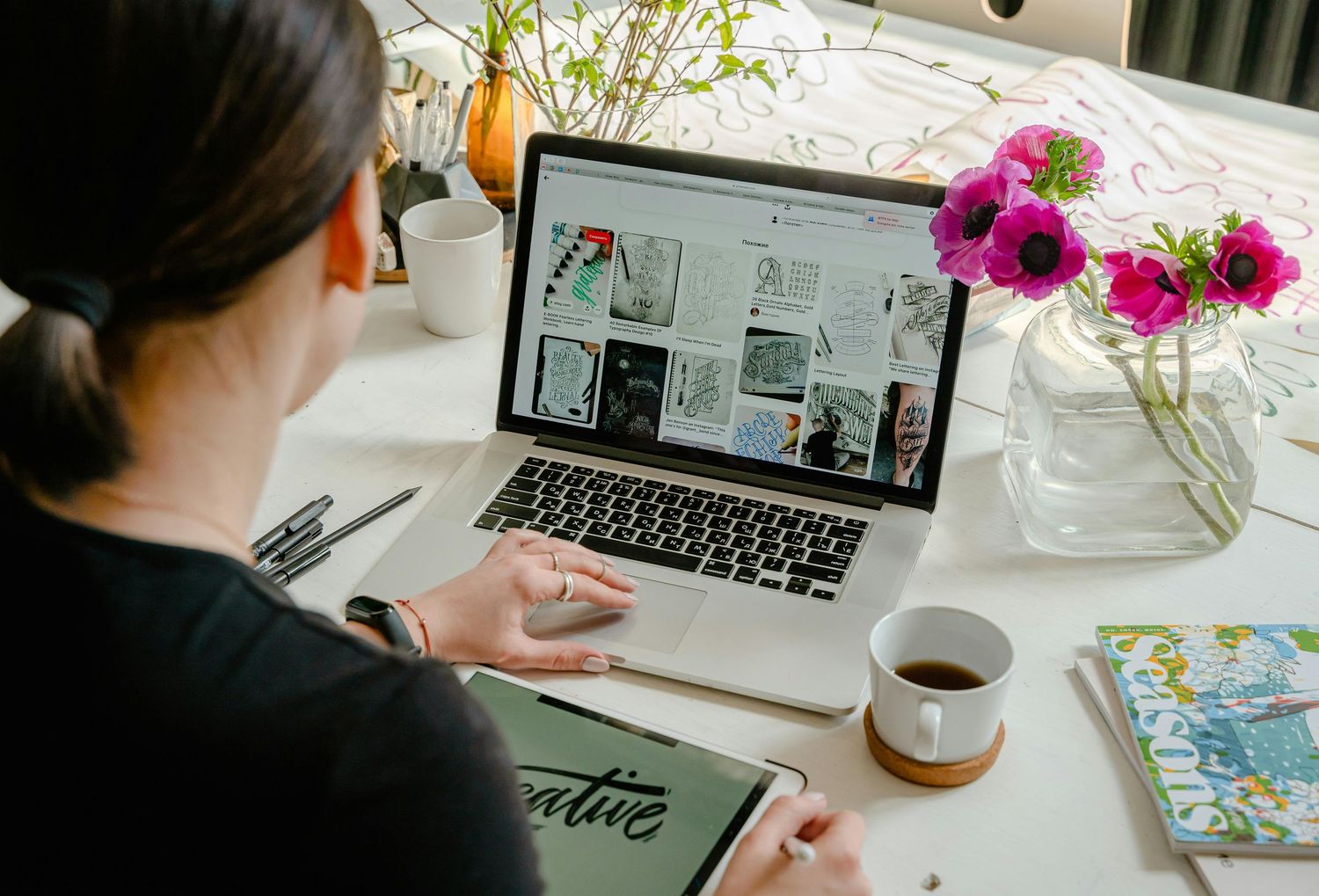 Person working at desk with laptop, coffee, and pink flowers in modern home office setting.