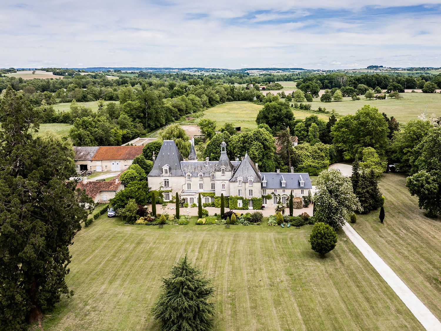 Aerial panoramic view of a luxurious estate with manicured lawns and tree-lined driveway in the countryside.