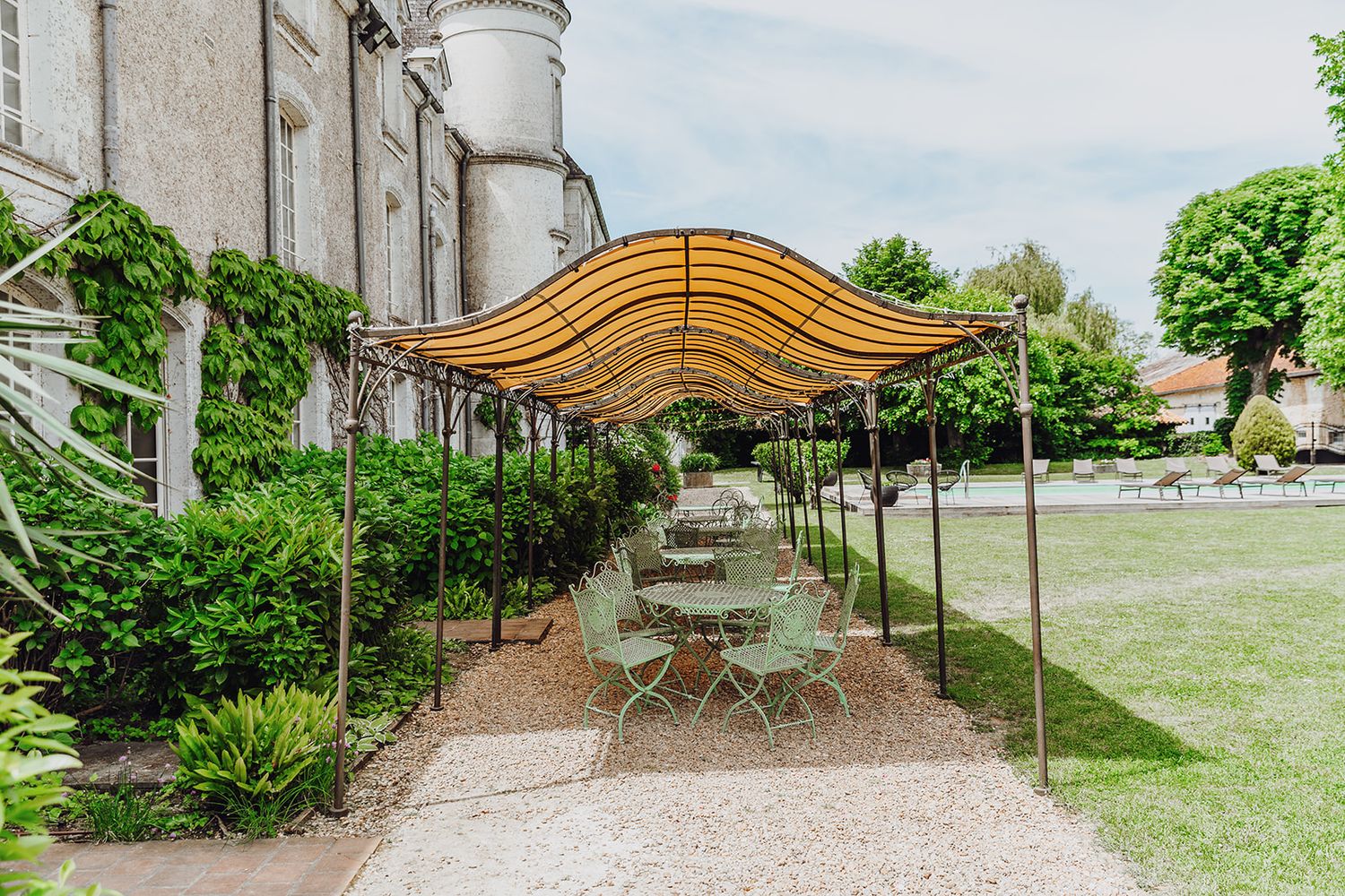 A curved wooden pergola structure creates a shaded walkway through a garden with seating areas and lush landscaping.