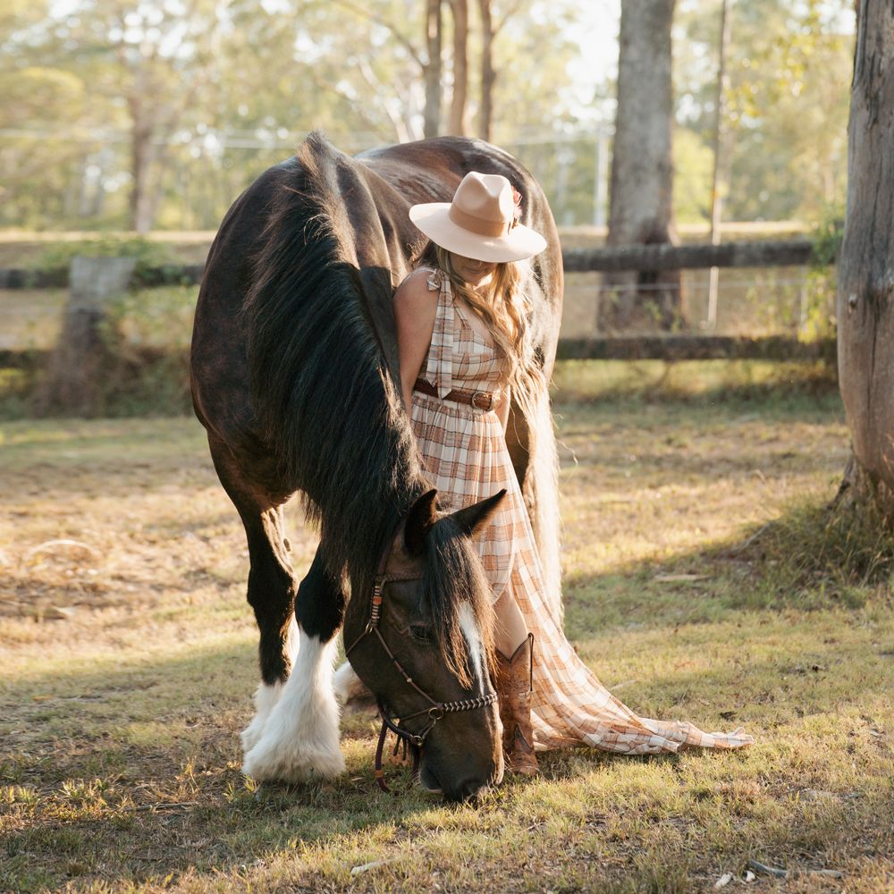 person in a flowing dress stands next to brown horse in a sunlit autumn field with wooden fencing during a photoshoot with a horse in Sydney