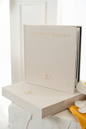 White wedding album with gold lettering displayed on white and yellow fabric background.