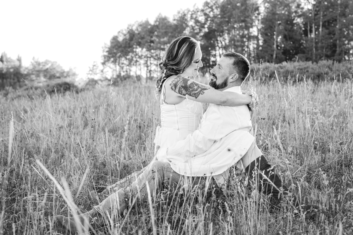 Black and white photo of a couple embracing and kissing in a meadow during an outdoor engagement session. She sits in his lap facing him.