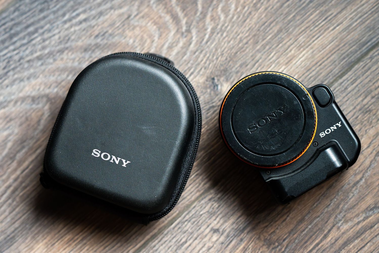 A black camera lens cap with a cord attached sits on a wooden surface next to its protective case.
