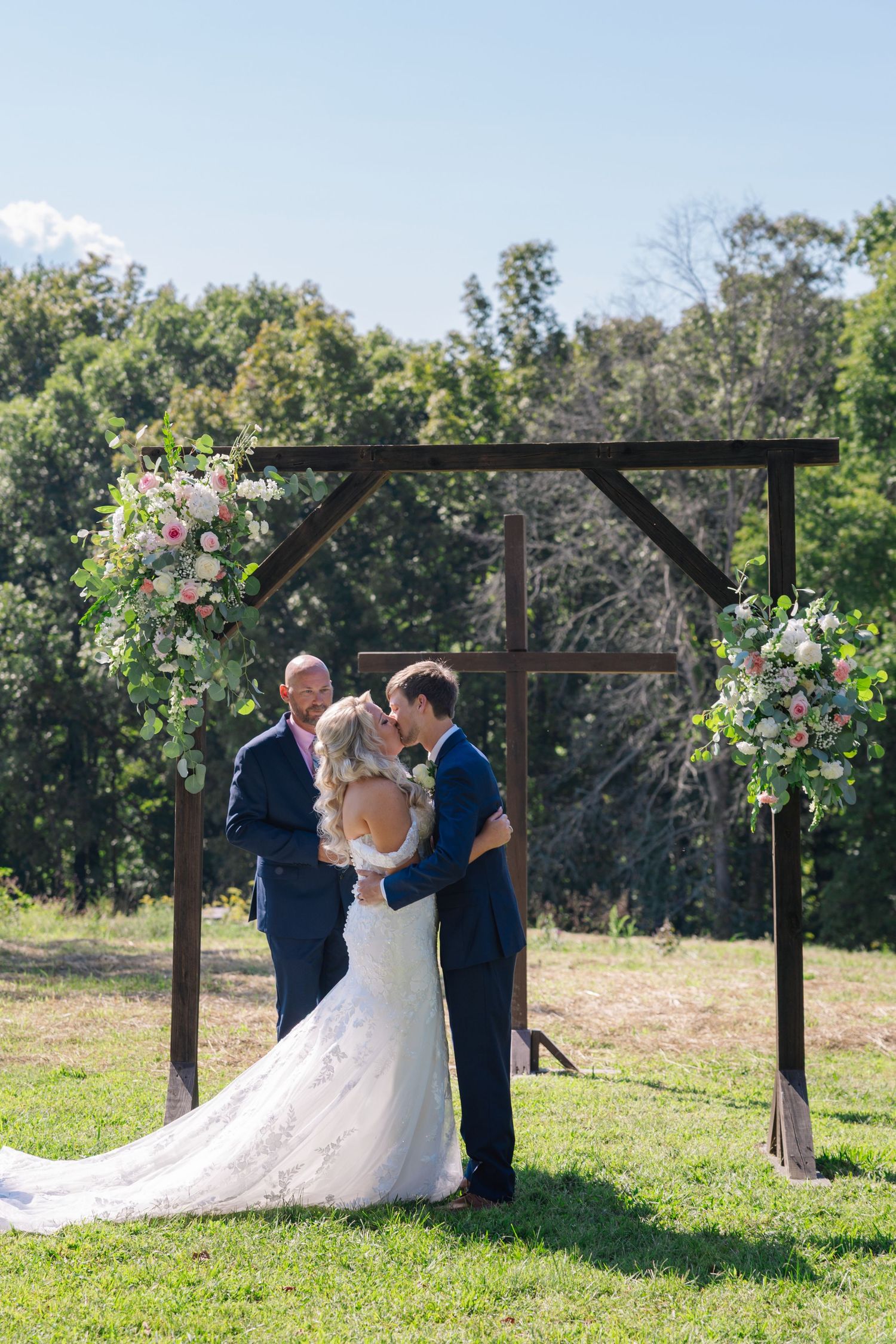 Outdoor wedding ceremony sequence under a wooden arbor decorated with pink flowers in a sunny field.