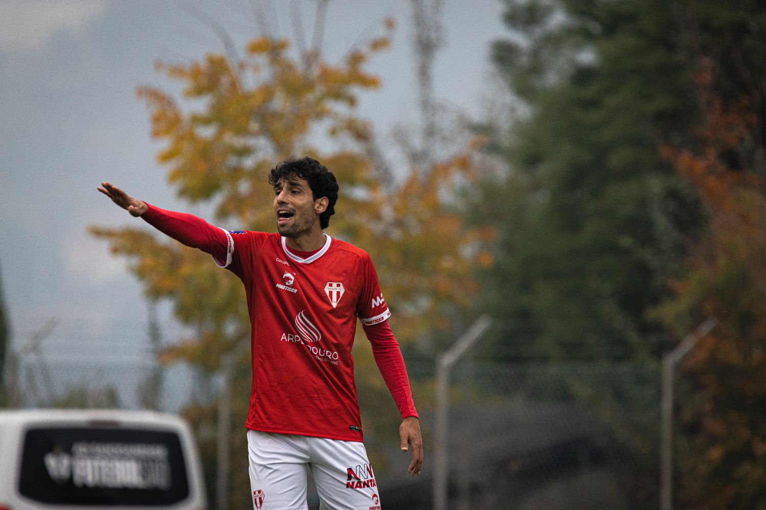 Soccer players in red and black uniforms compete during an autumn match with trees in background.