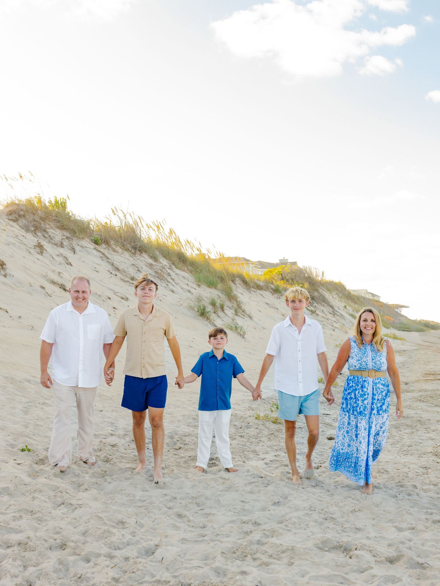 Family group walking and holding hands along a sandy beach dune in coordinated blue and white summer outfits.