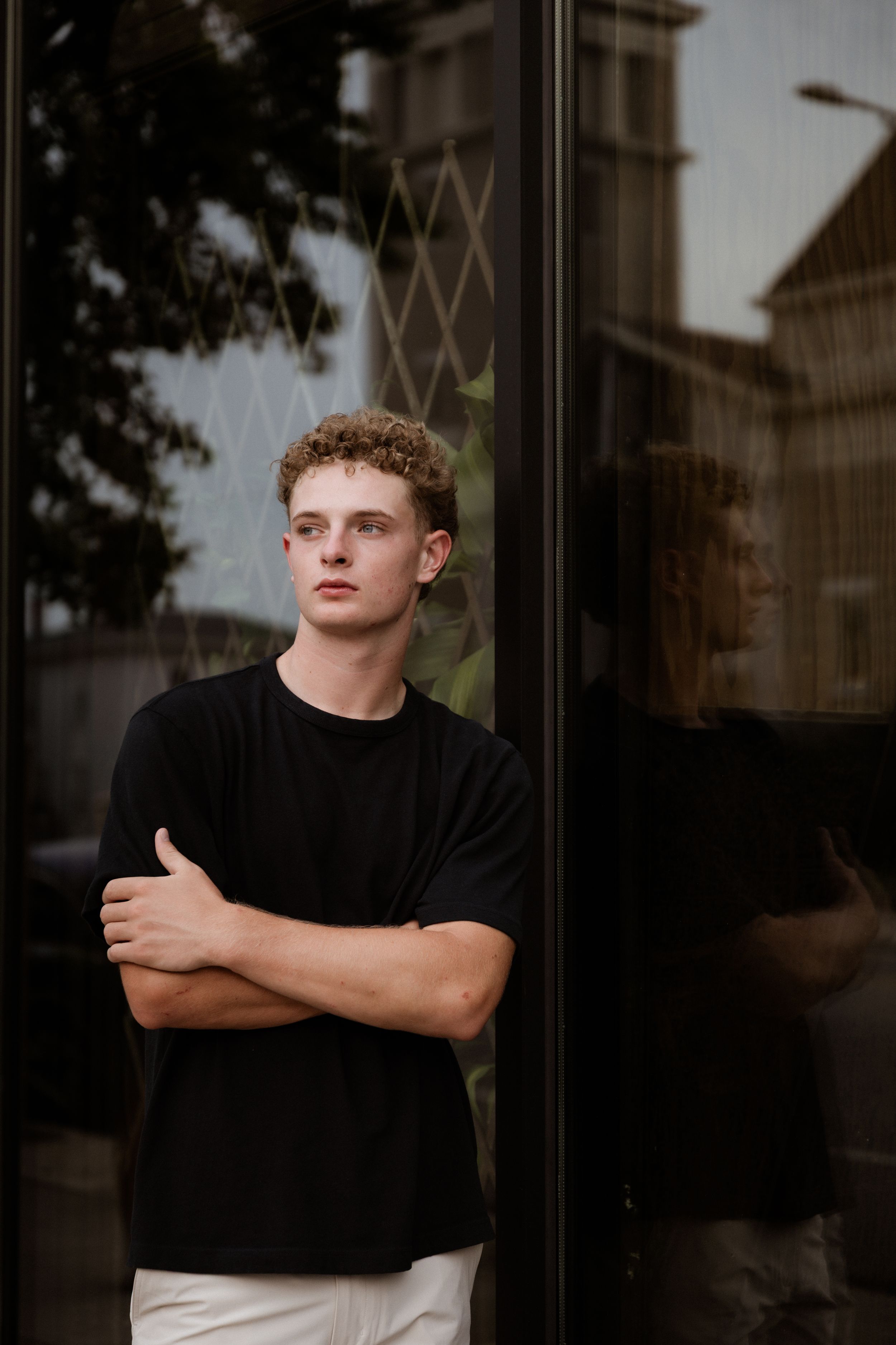 Young man with curly hair in black t-shirt stands arms crossed against glass building exterior.