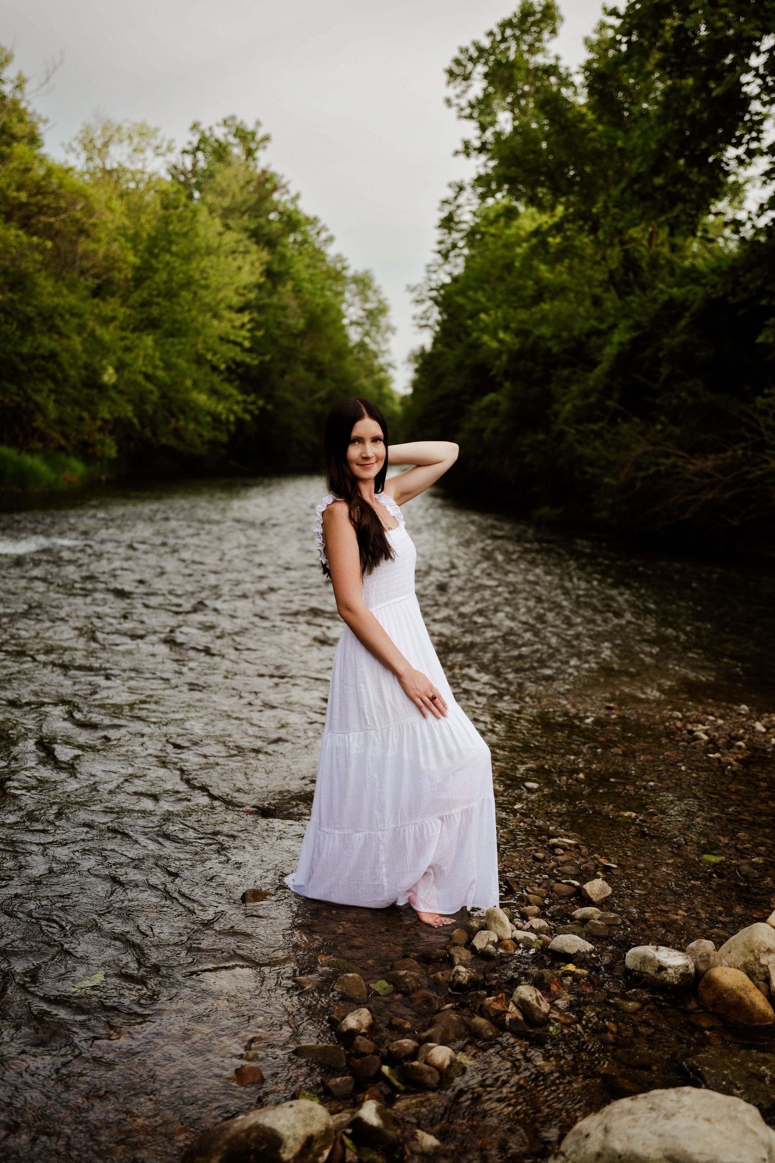 Brunette woman in flowing white maxi dress standing in rocky river surrounded by green trees.