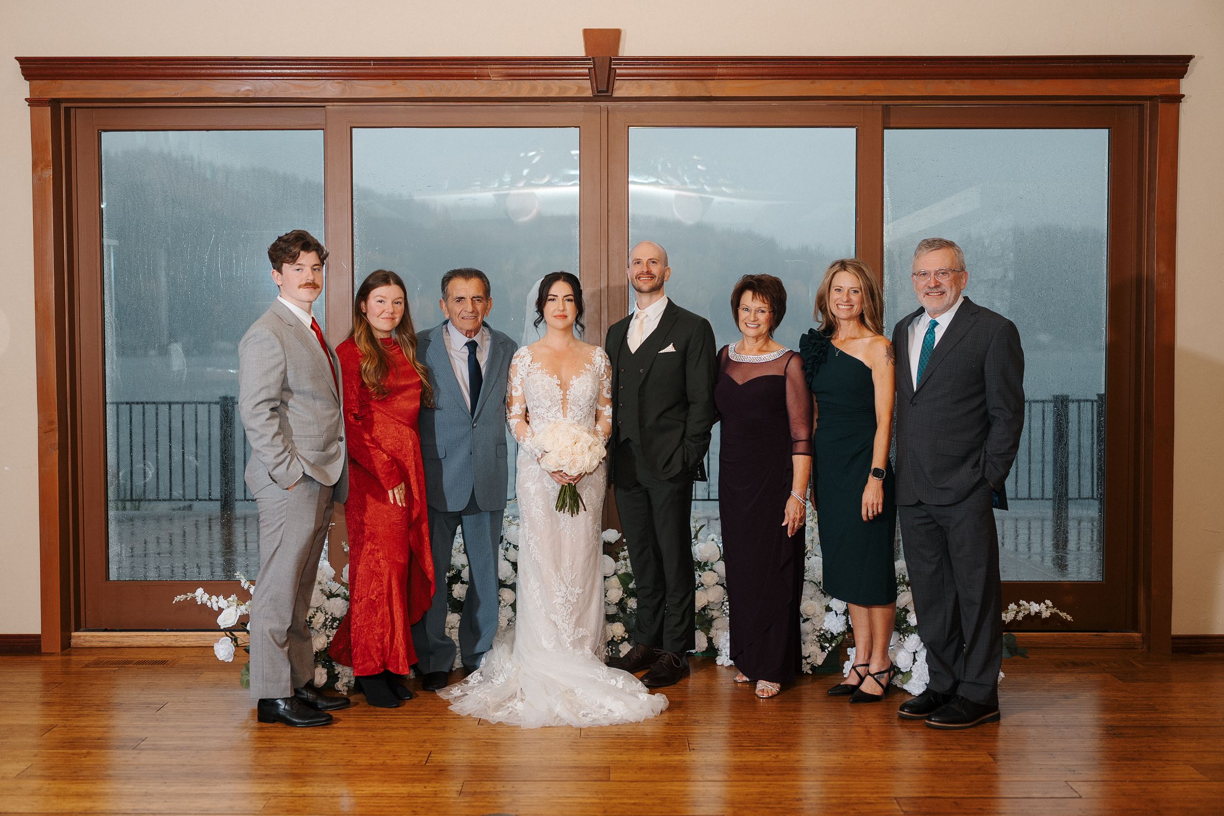 Wedding party members lined up in formal attire against a wooden wall with windows overlooking water at sunset.