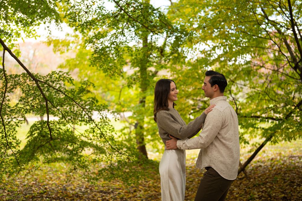 Romantic couple holding hands and gazing at each other under lush green autumn trees in a park.