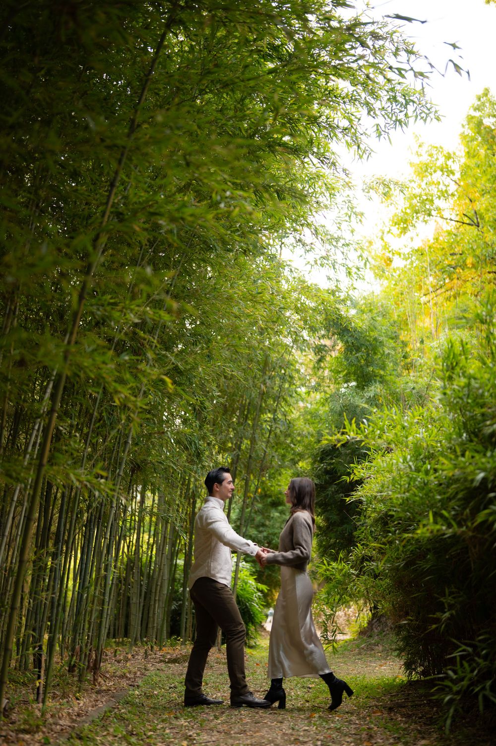Couple holding hands facing each other on a serene bamboo forest pathway surrounded by tall green bamboo.