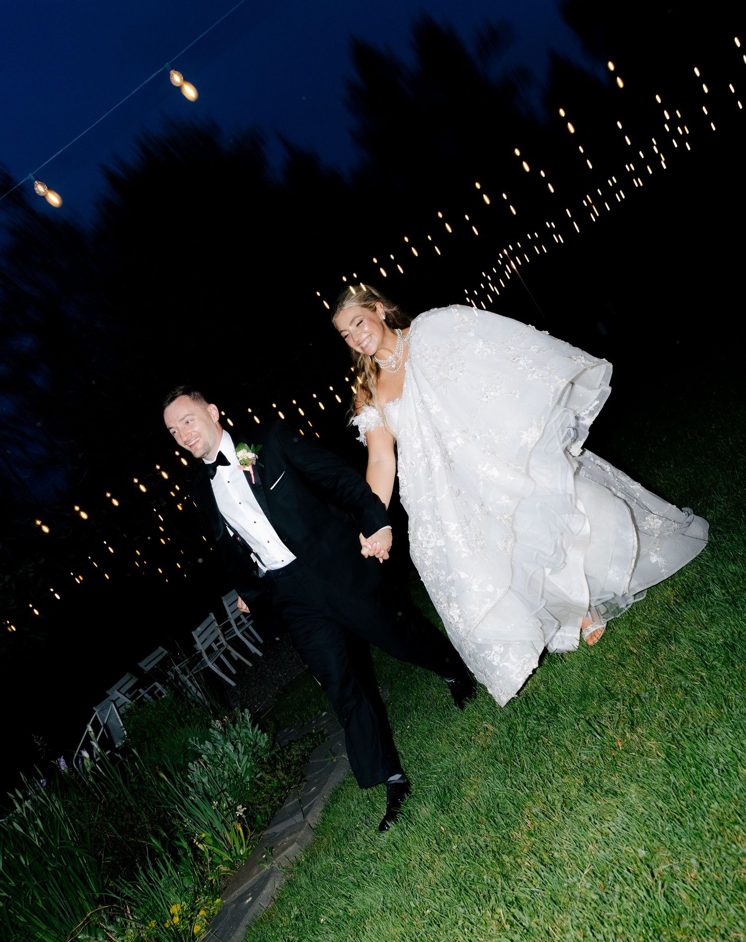 A couple in formal attire dips dramatically on a grassy lawn under twinkling string lights at night.
