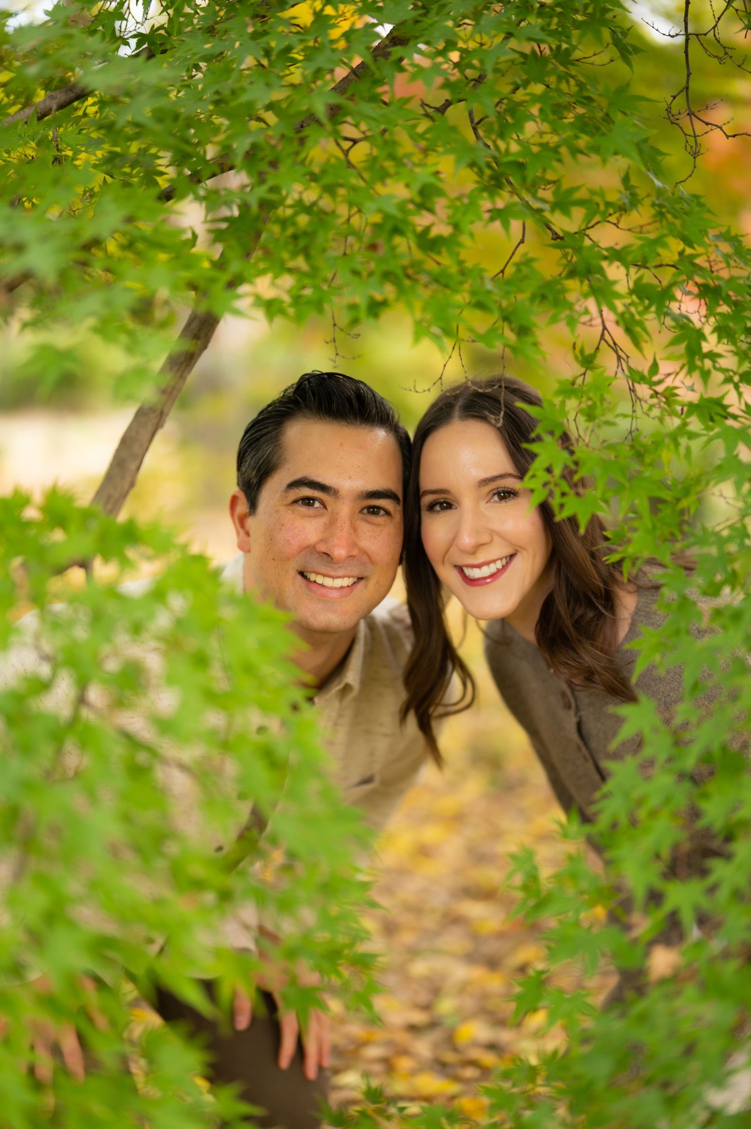 Happy couple smiling through lush green maple tree branches in a vibrant autumn garden.