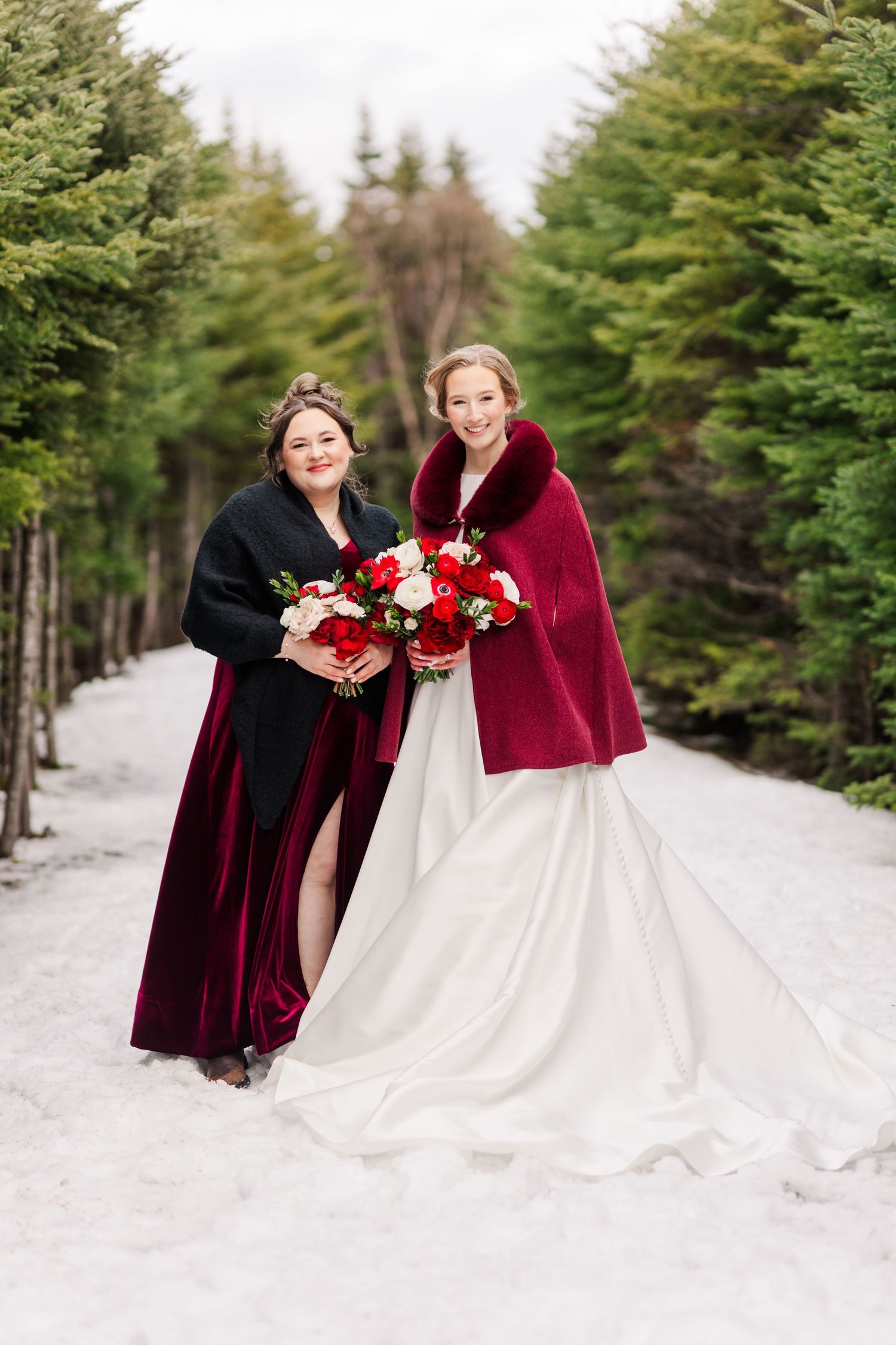 Winter wedding party wearing burgundy velvet wraps pose together in snowy evergreen forest setting.