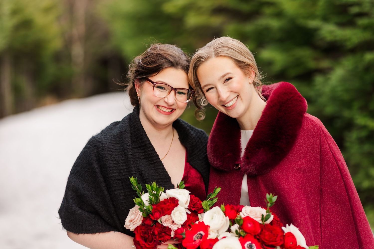 A couple in burgundy and black attire embraces in a snowy winter forest setting with evergreen trees.