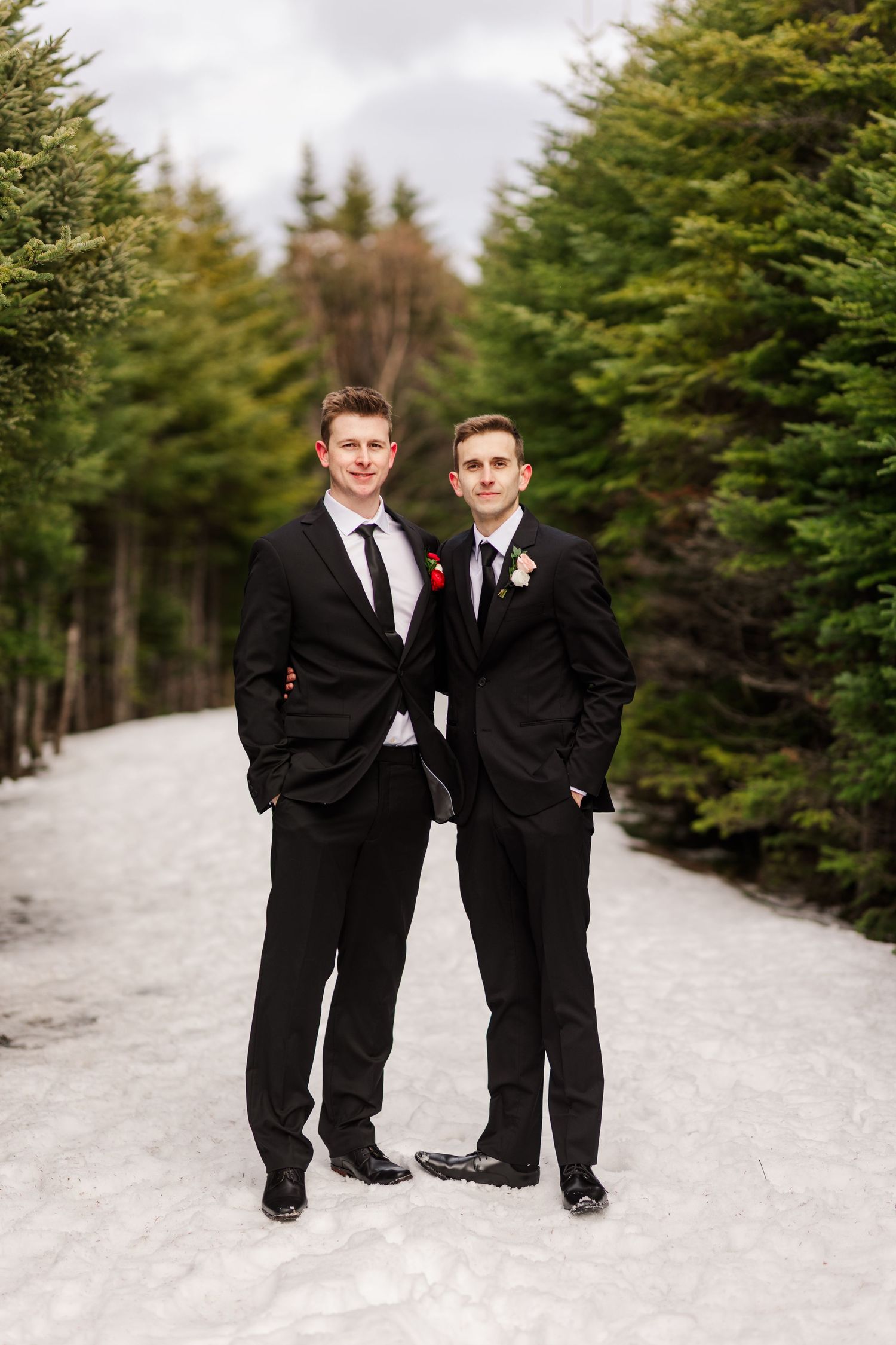 Two people in black suits pose together on a snowy path lined with evergreen trees in winter.