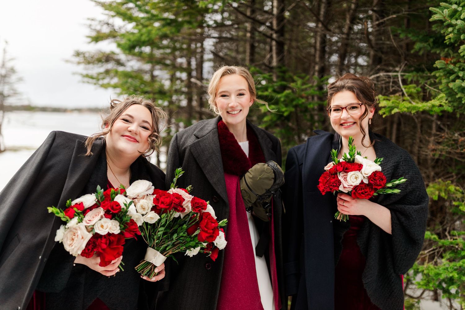 Wedding party laughs together while holding red and white floral bouquets in an outdoor winter setting.