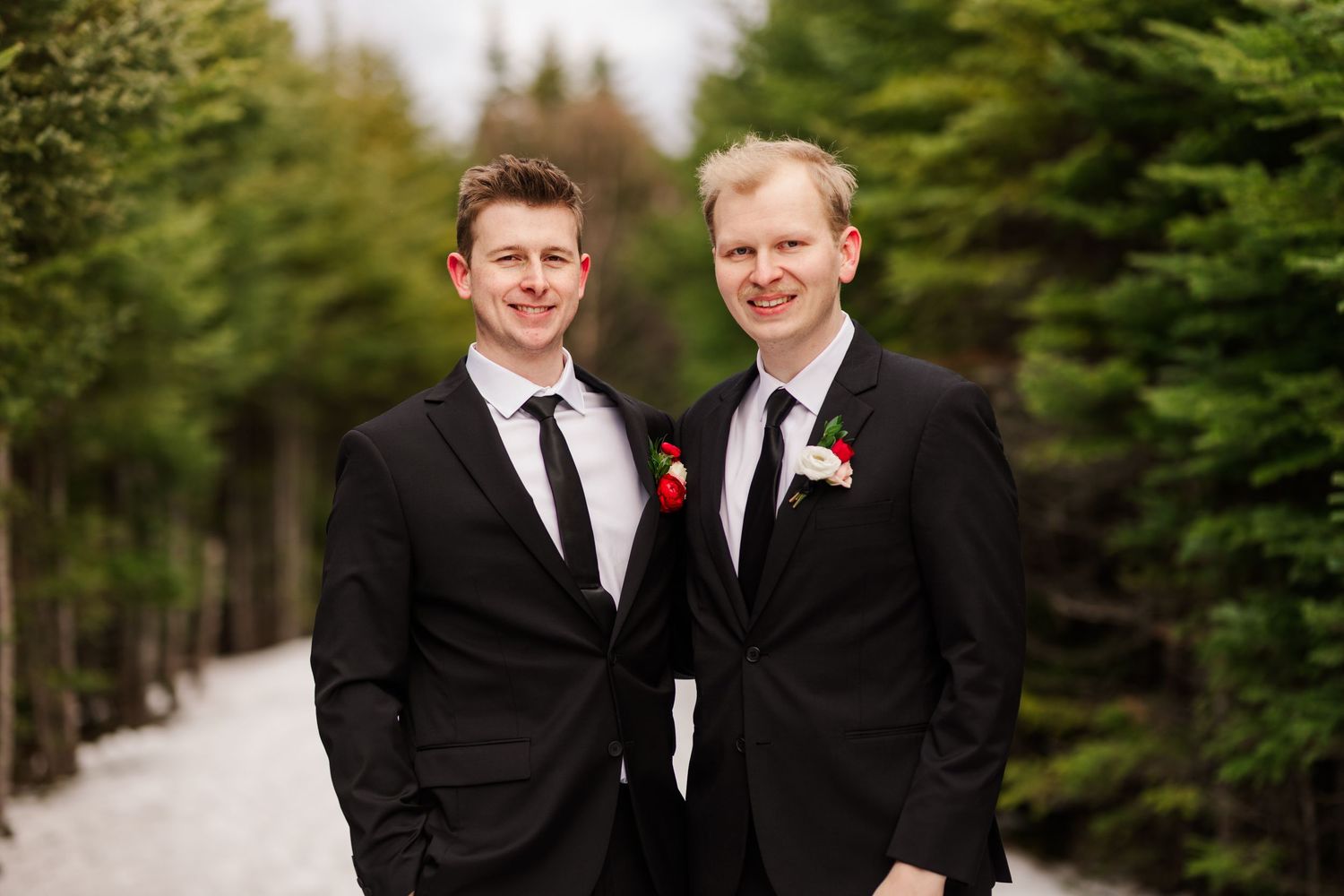 A wedding party in formal black suits and ties poses together against a backdrop of snow-covered evergreens.