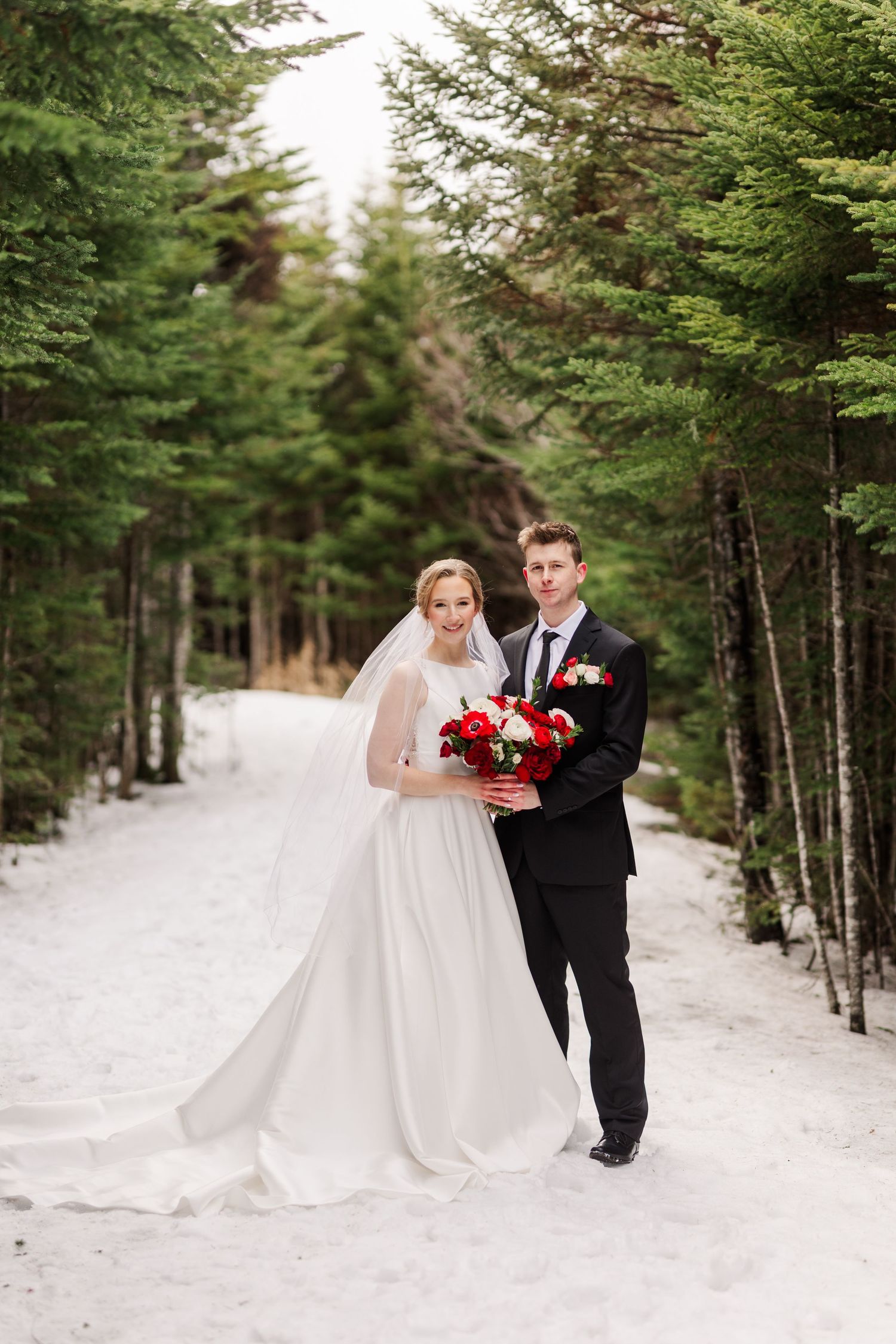 A winter wedding couple poses in a snowy evergreen forest path wearing traditional formal attire.