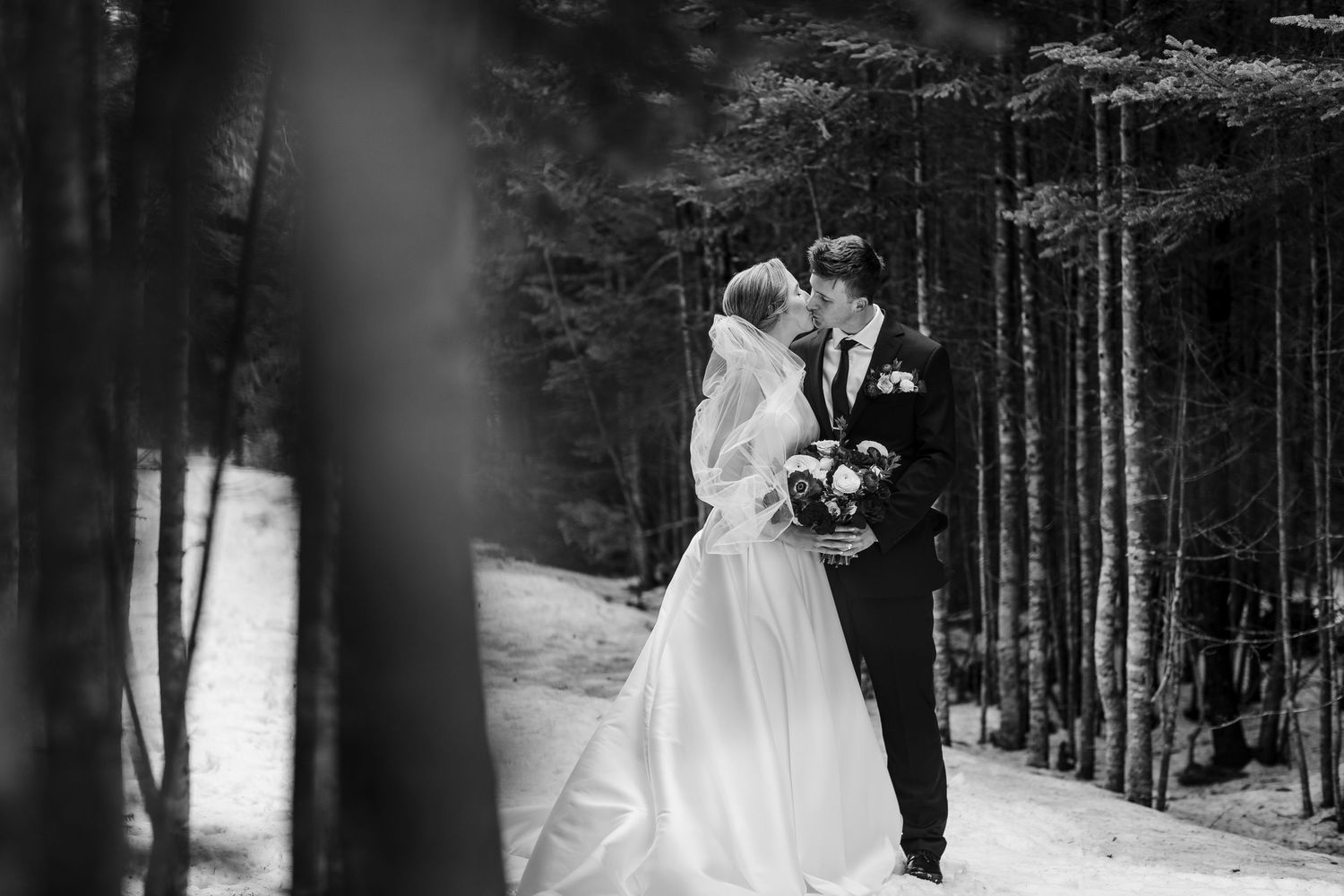 A couple in wedding attire embraces in a snowy winter forest setting during their wedding photoshoot.