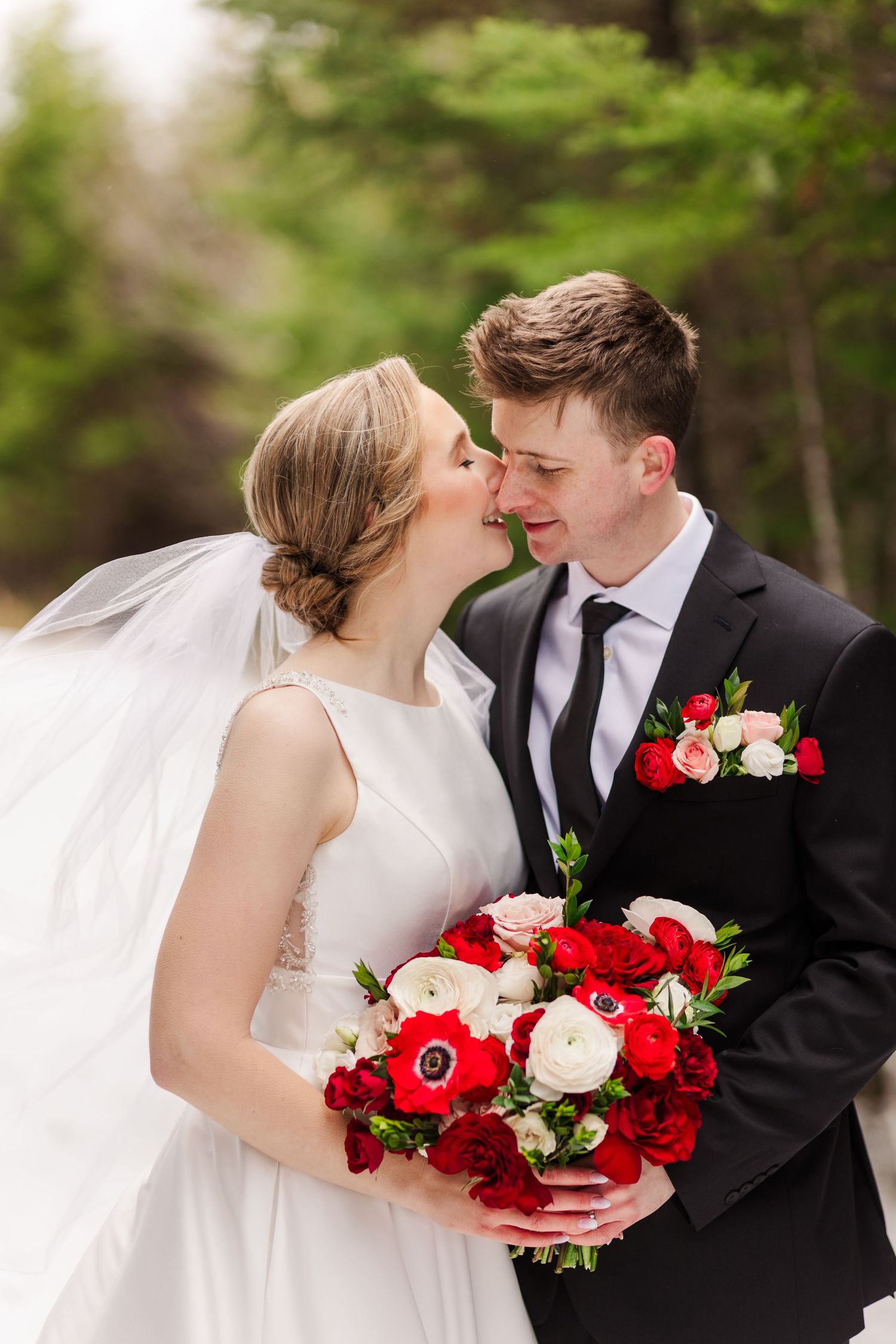A romantic wedding couple shares tender moments with red and white floral bouquet in an outdoor garden setting.
