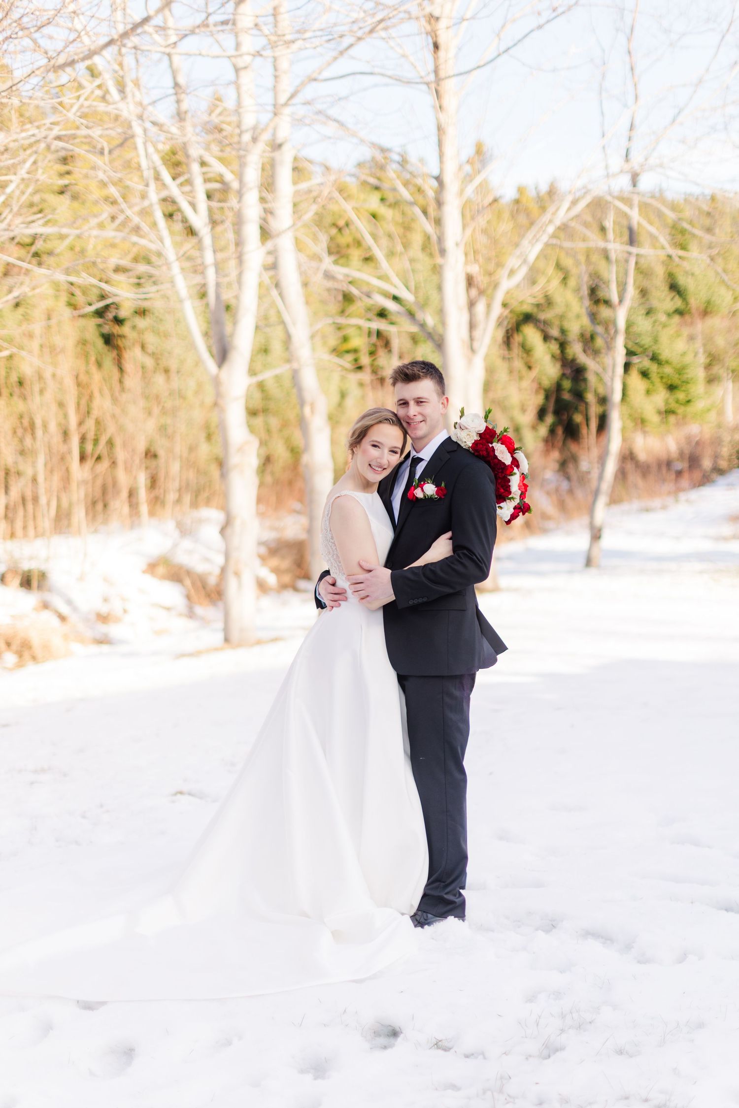 A couple in wedding attire shares a romantic moment in a snowy winter landscape with bare trees in the background.