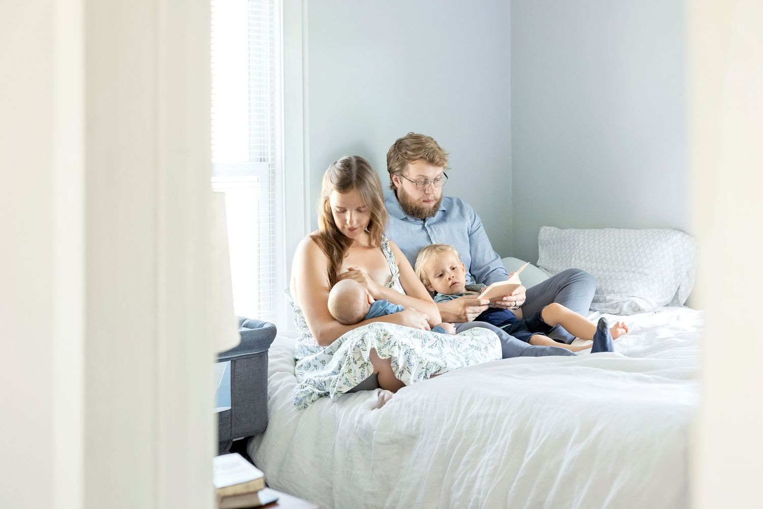 A Garner, Iowa family relaxes together on a white bed while reading a book to their newborn boy and toddler.