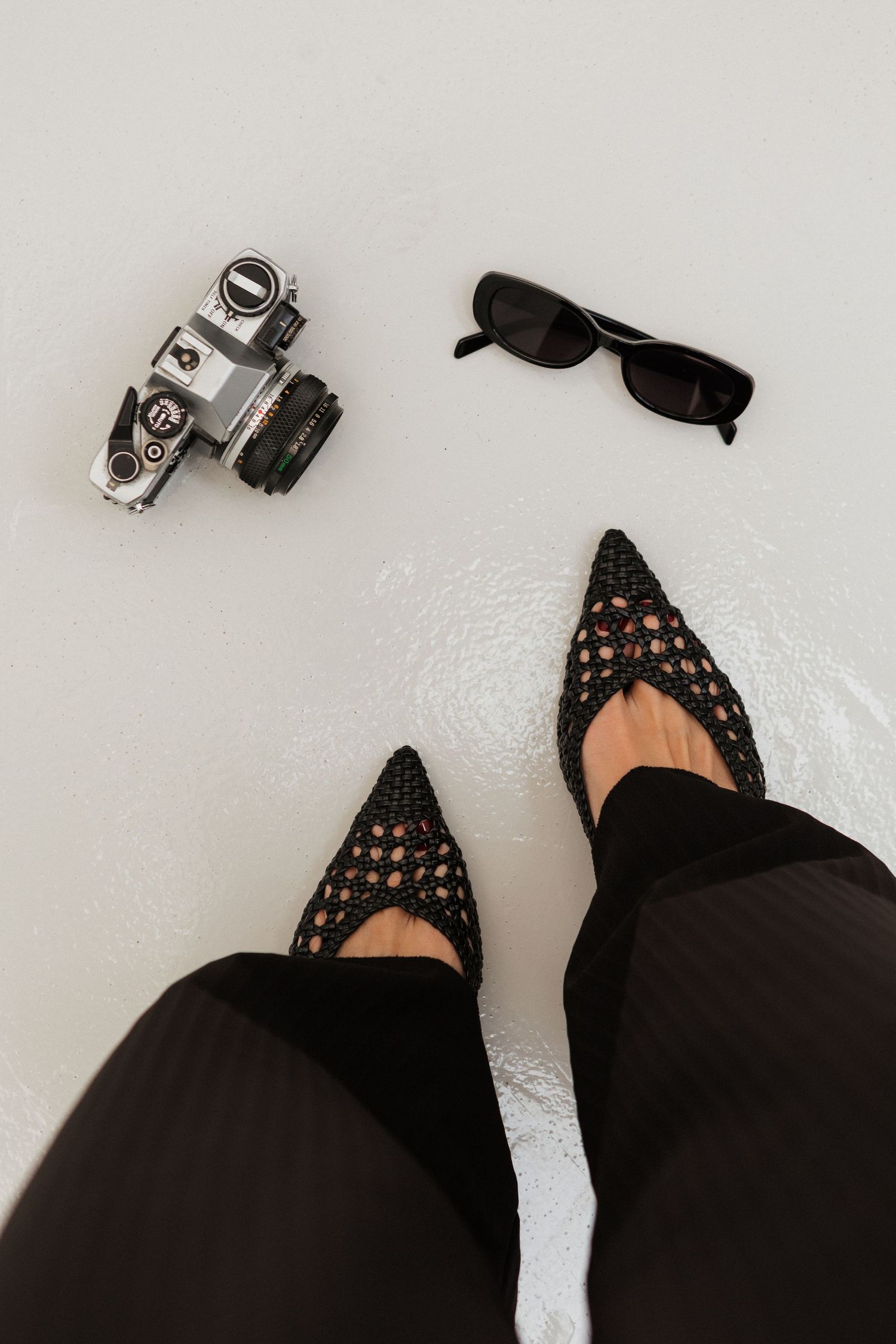 Lux black studded ballet flats with pointed toes paired with black pants on a white surface. Editorial style image with vintage camera.