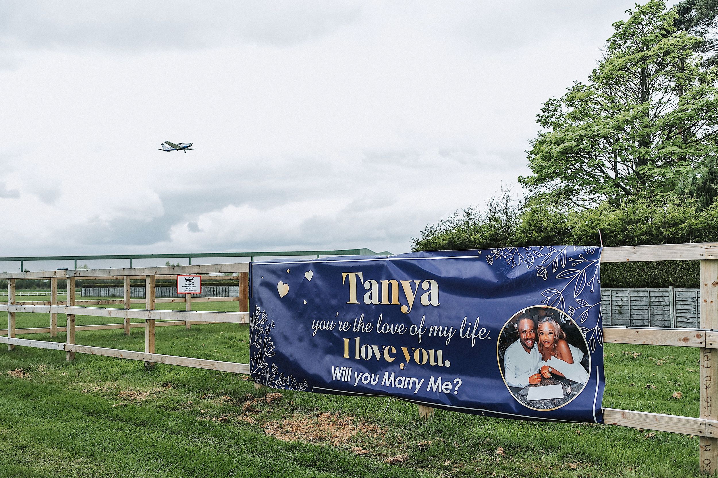 A series of outdoor banners displayed along a wooden fence in a rural setting with green trees in the background.