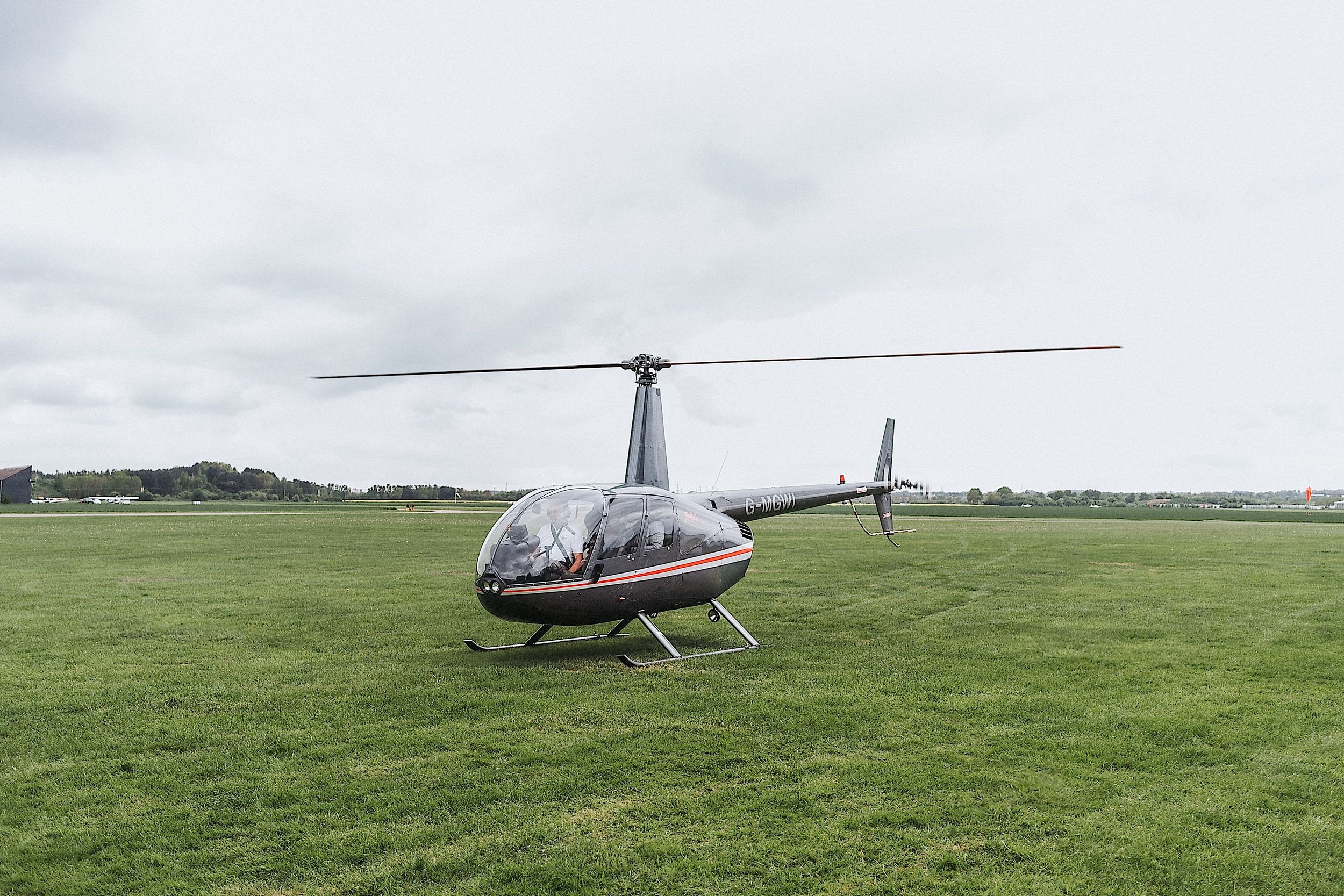 A red and white Robinson R44 helicopter lands in a grassy field on a cloudy day.