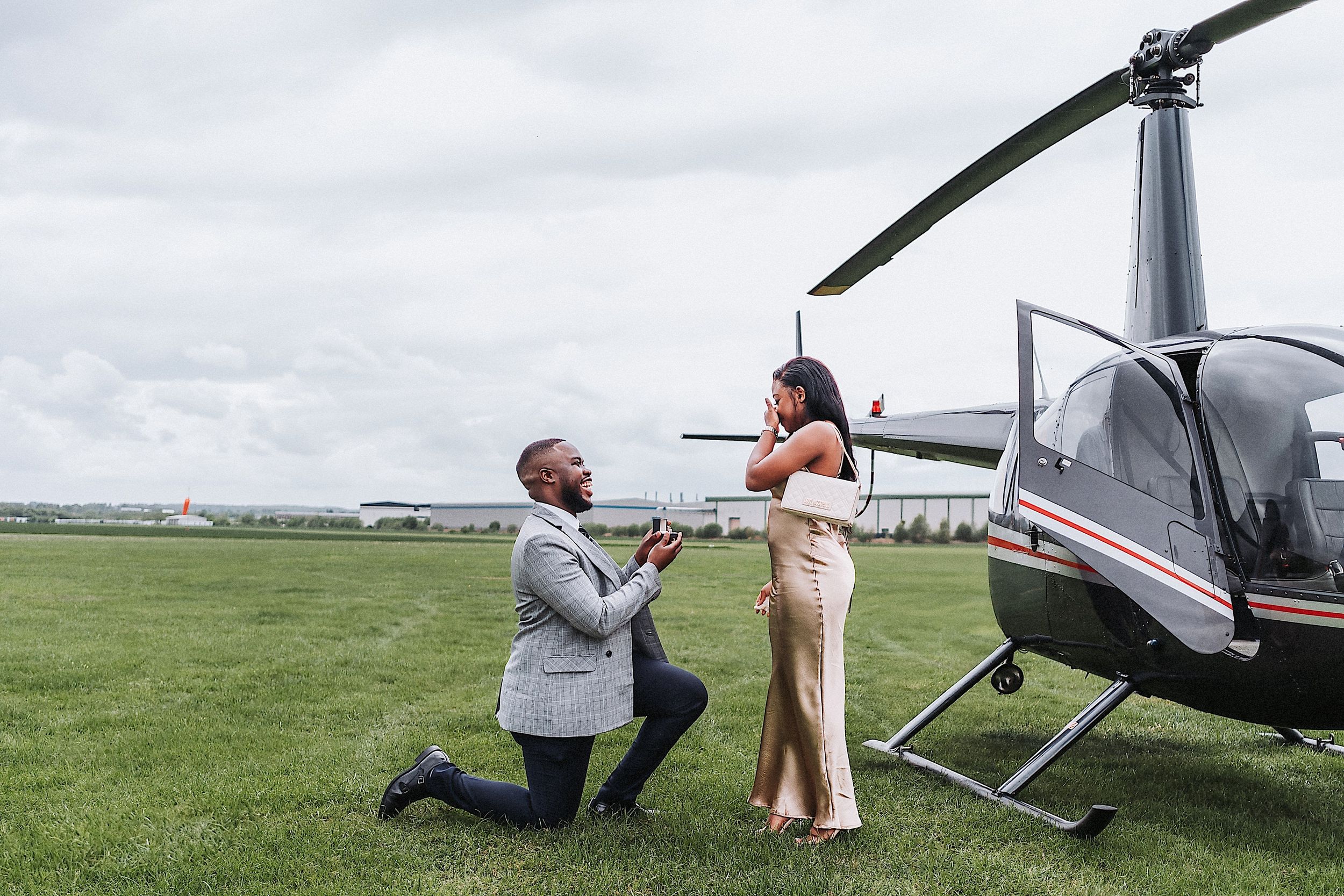 A romantic proposal takes place in front of a small aircraft on a grassy airfield on a cloudy day.