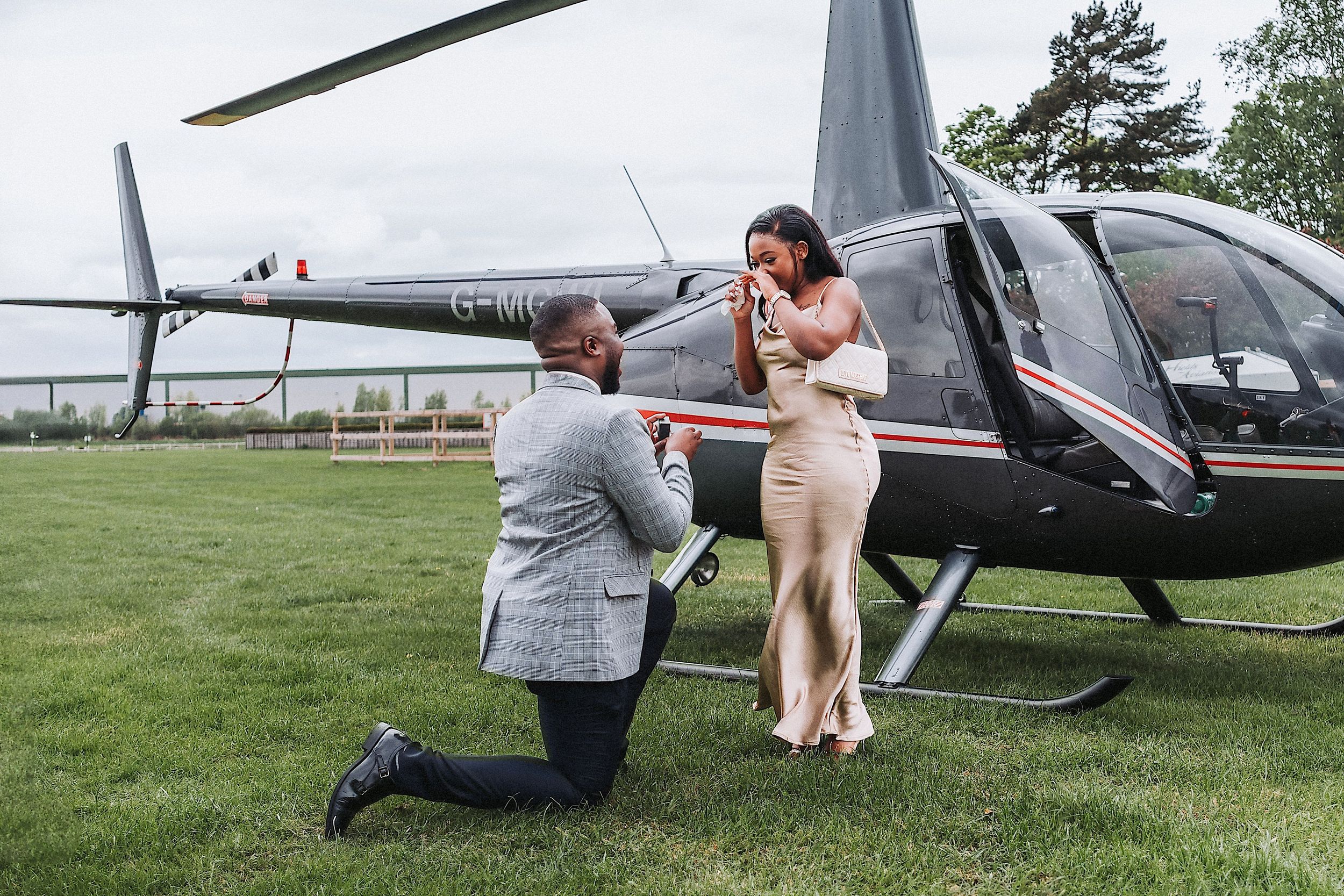 A romantic helicopter proposal scene unfolds on a grassy field with a black helicopter in the background.