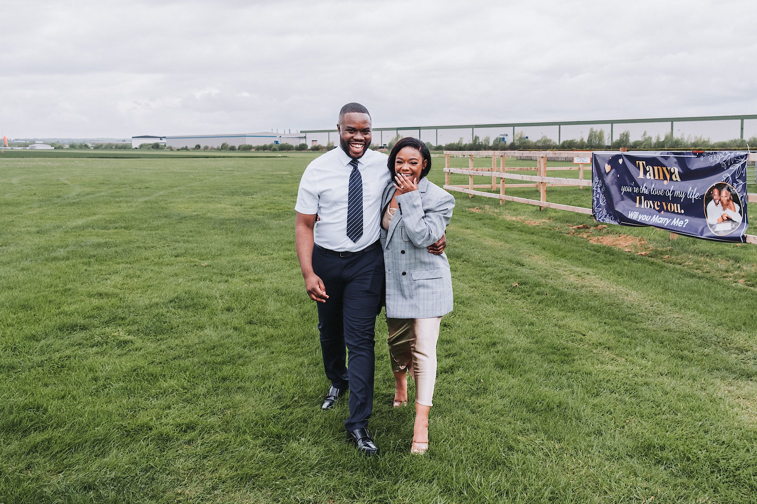 A couple shares a joyful moment together on a sunny day in a grassy field with wooden fencing in the background.
