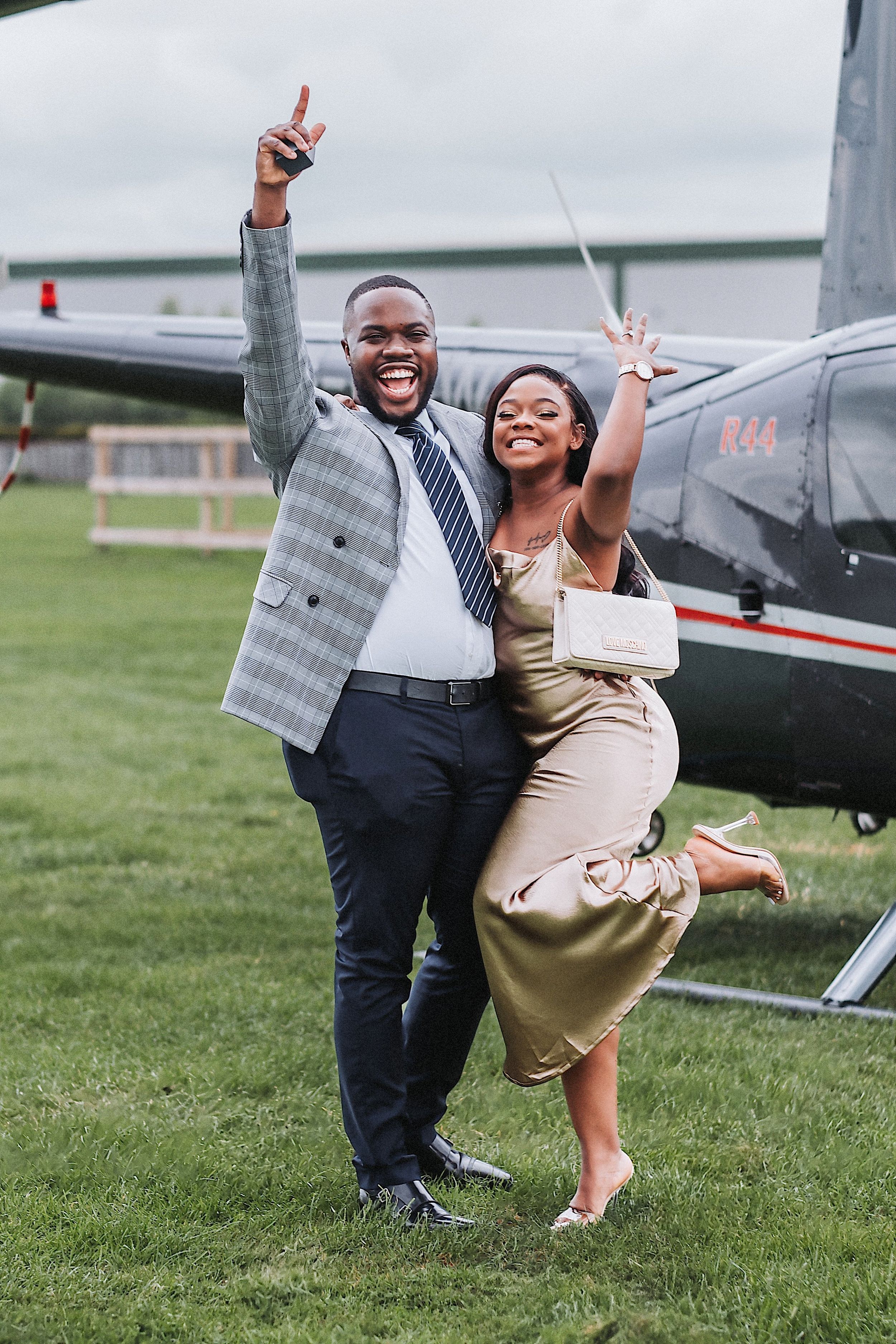 A joyful couple poses playfully on a grassy field next to a small aircraft on a sunny day.
