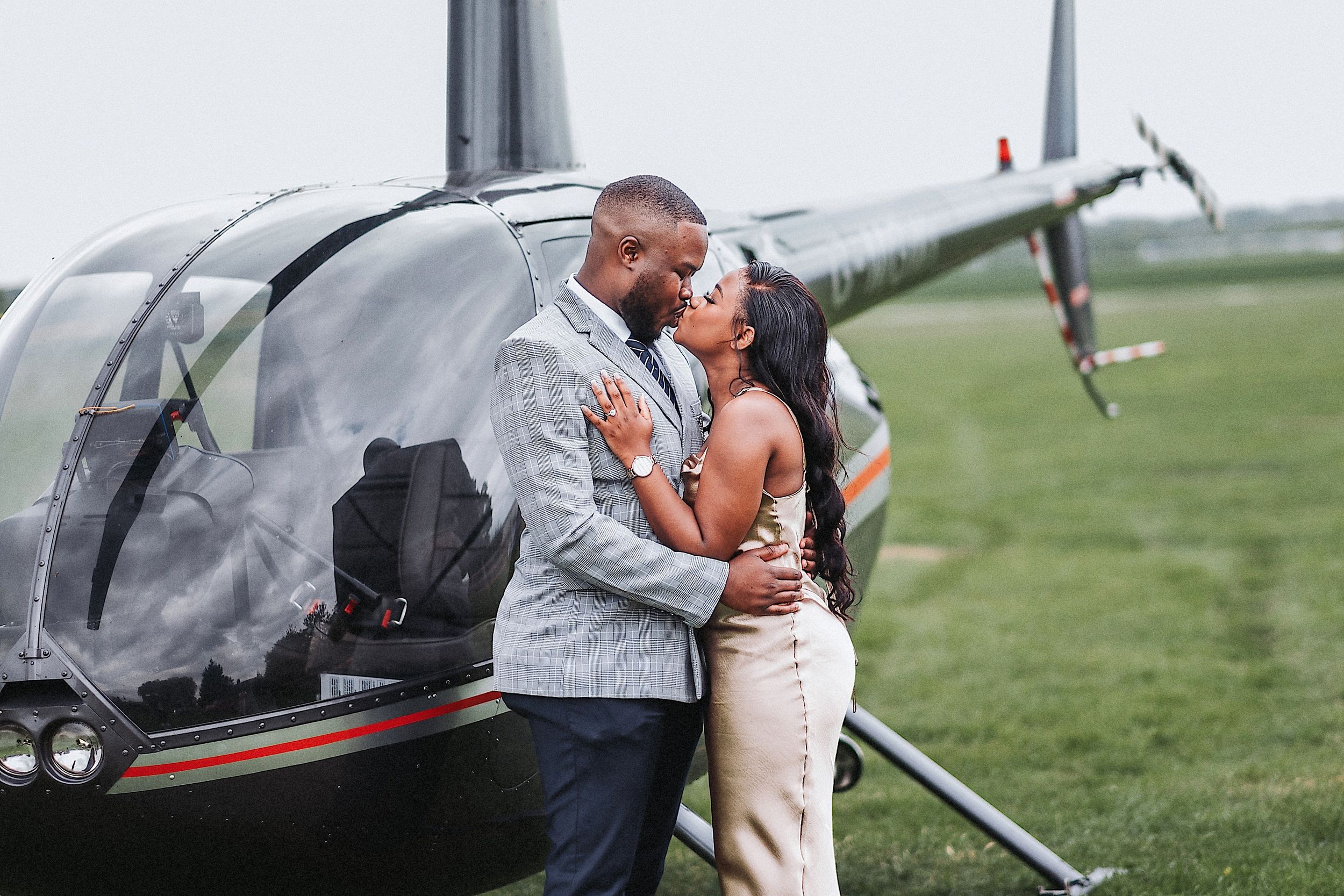 Romantic couple embraces and kisses near small private aircraft on grassy airfield.