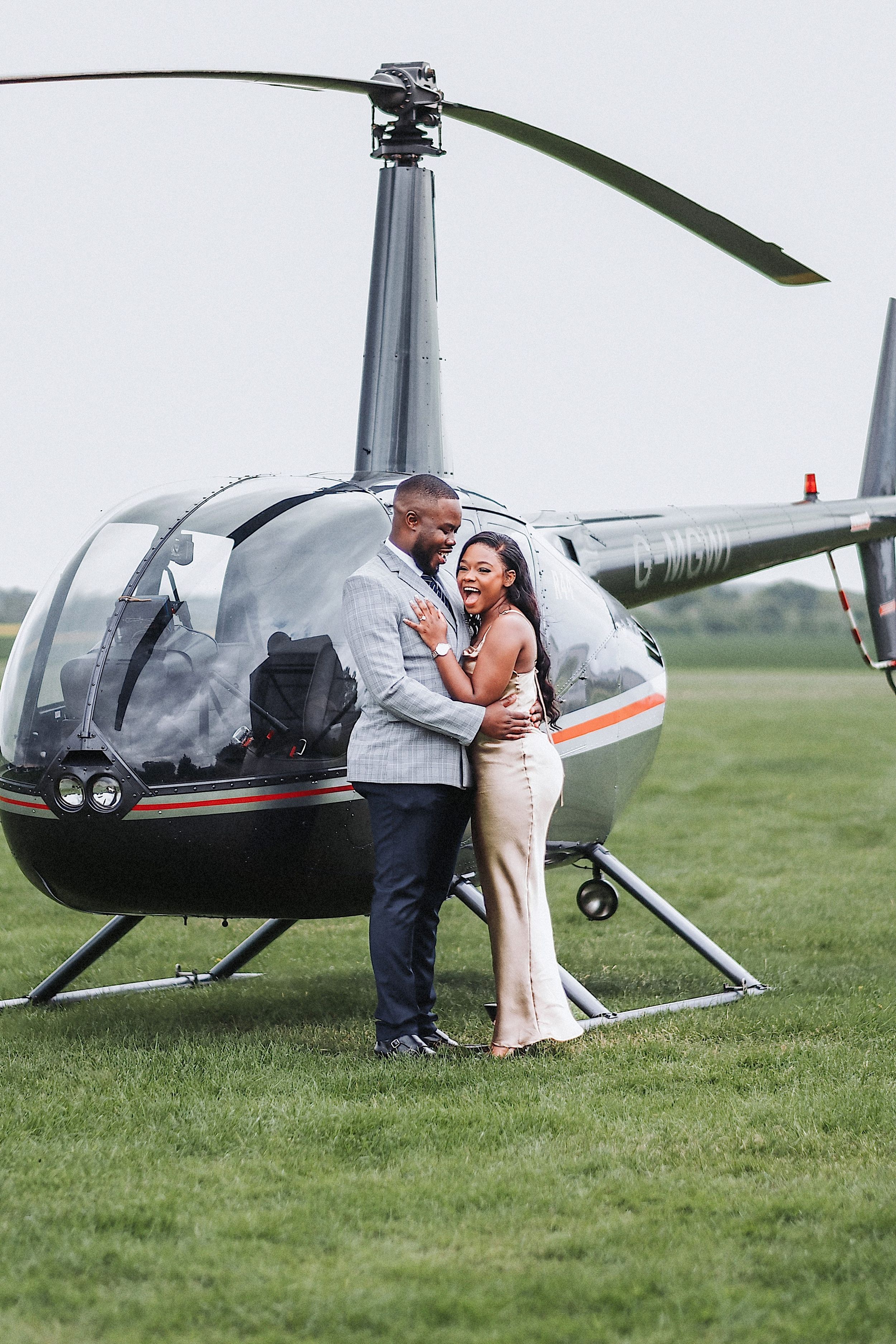 A couple shares a romantic moment next to a parked black helicopter on a grassy field.