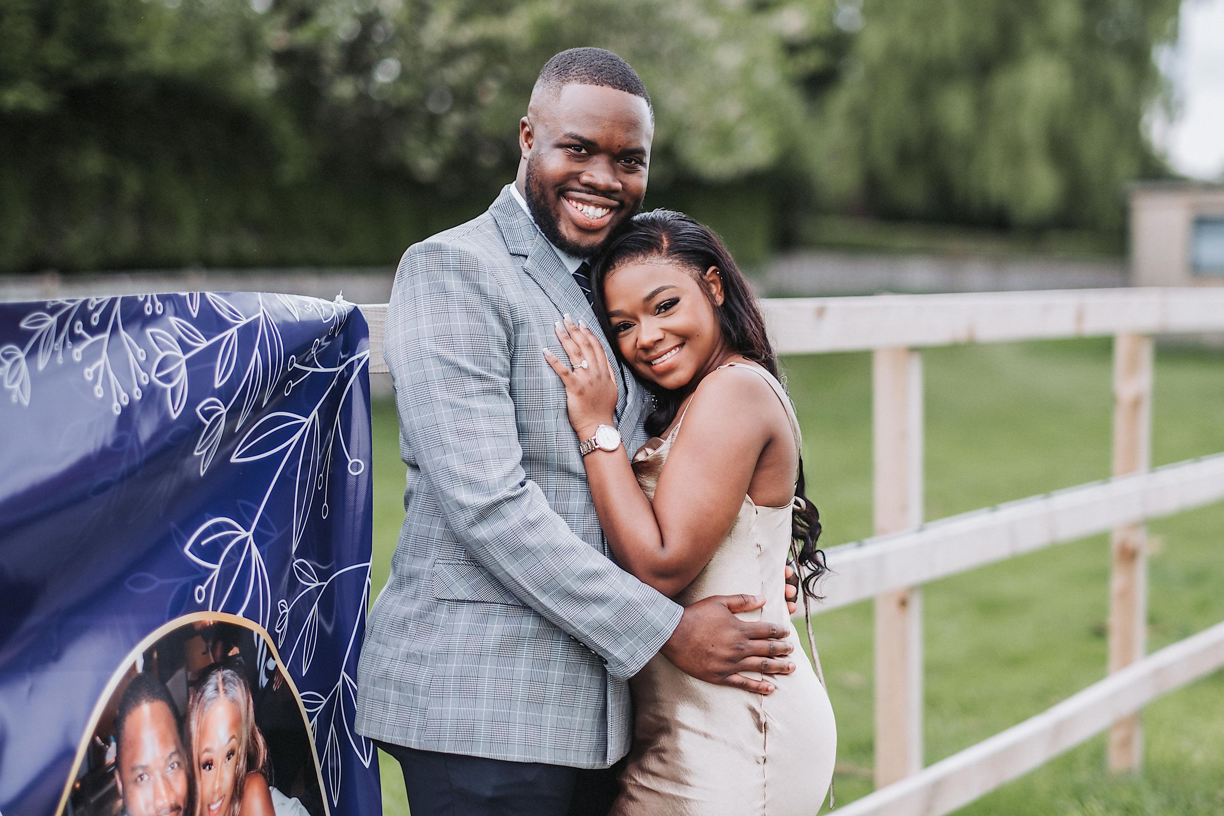 A joyful couple embraces and laughs together during an outdoor photoshoot near white fences on a sunny day.