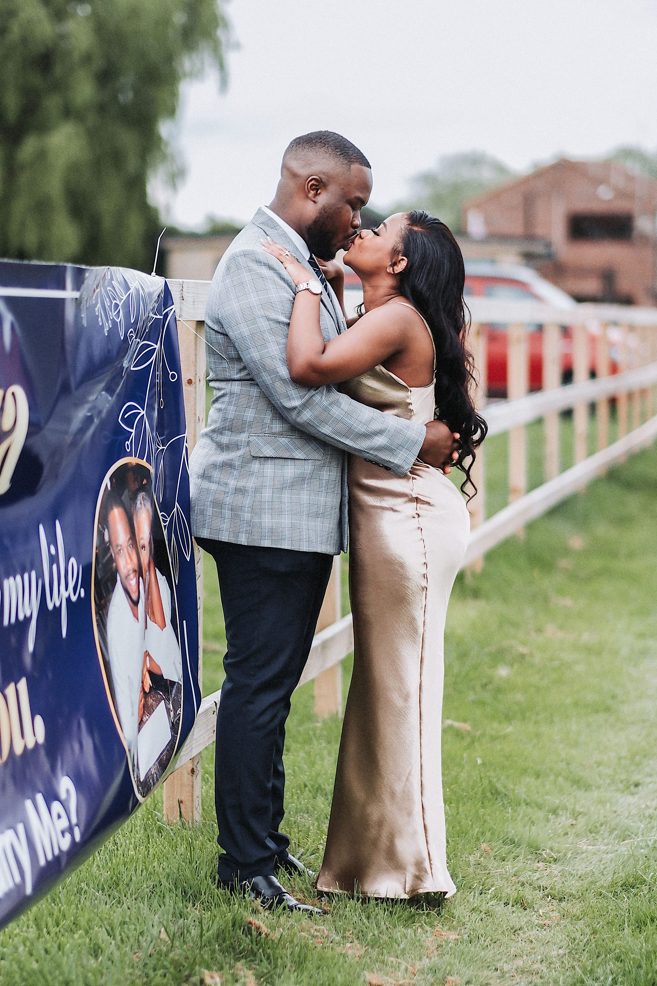A romantic couple shares tender kisses while leaning against a blue banner at an outdoor venue.