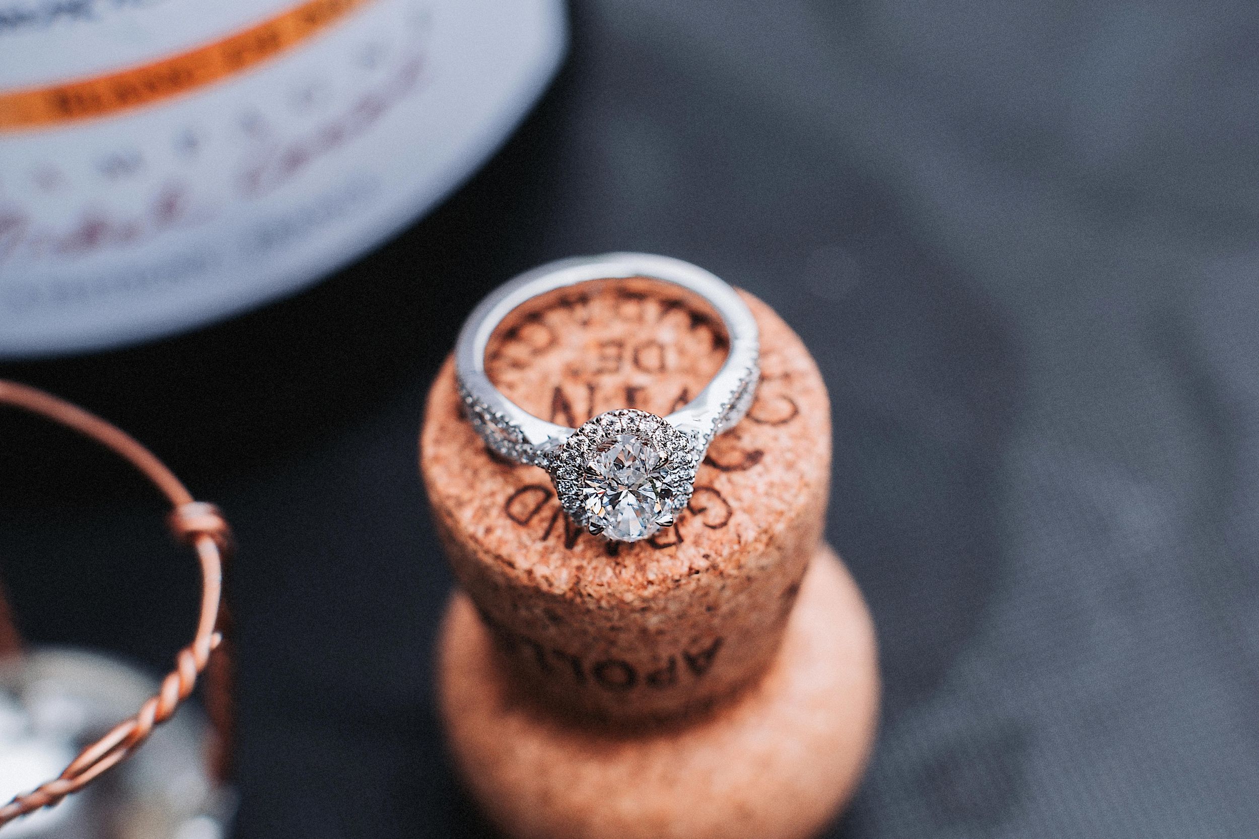 A diamond engagement ring displayed on a rustic cork bottle stopper against a dark background.
