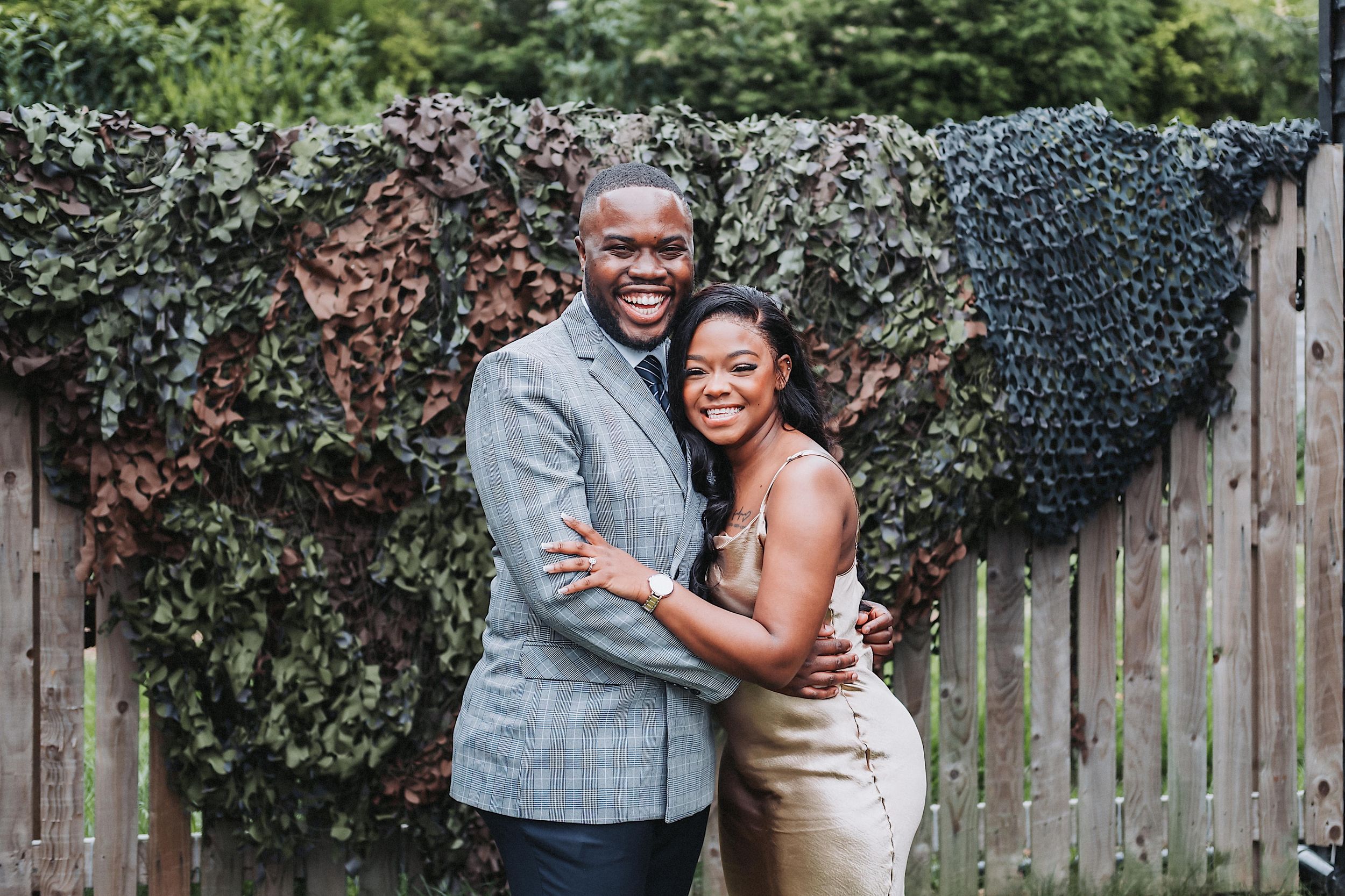 A couple shares joyful embraces and laughter in a backyard setting with wooden fence and greenery.