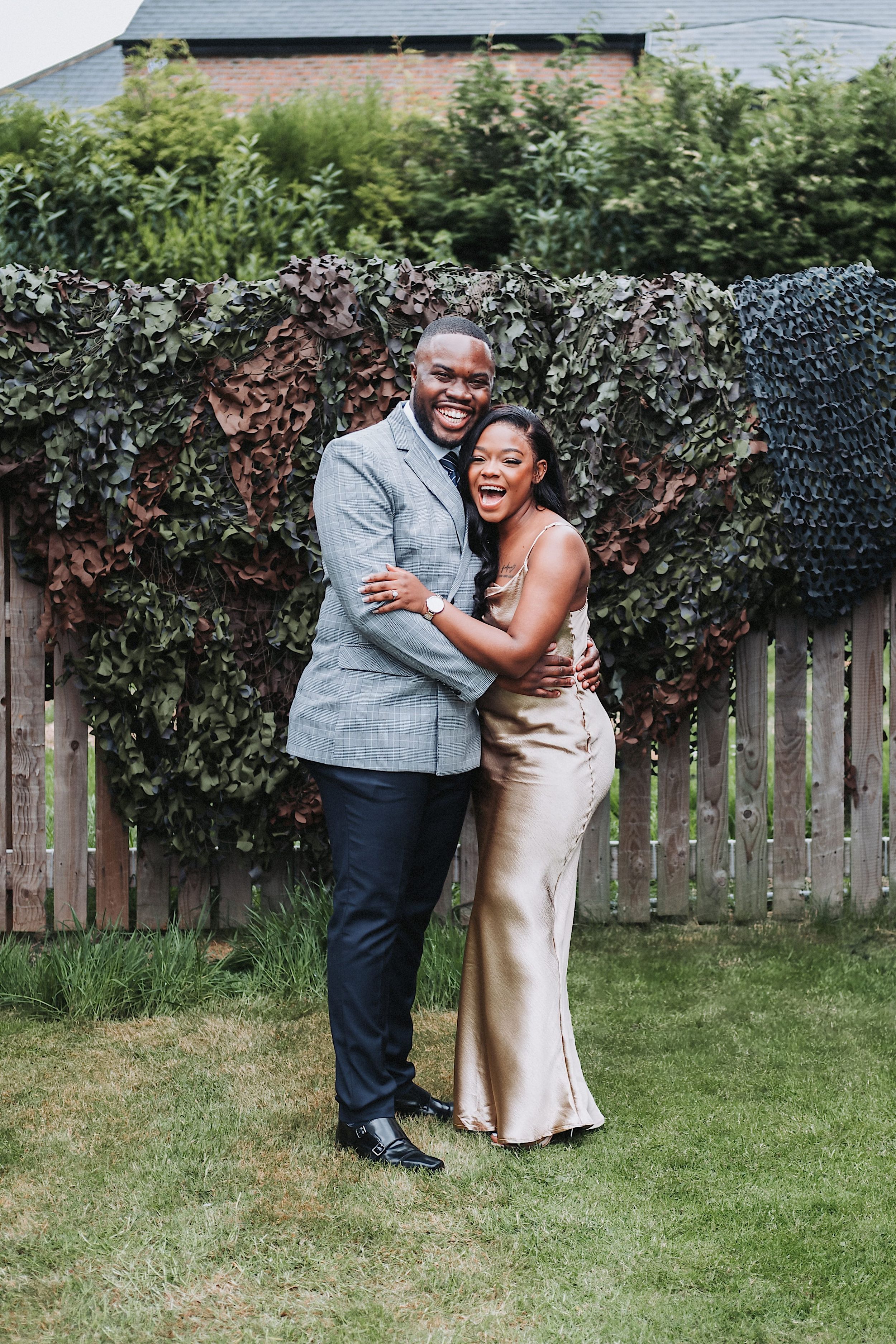 A couple shares a joyful embrace while standing in a backyard garden with a vine-covered fence behind them.